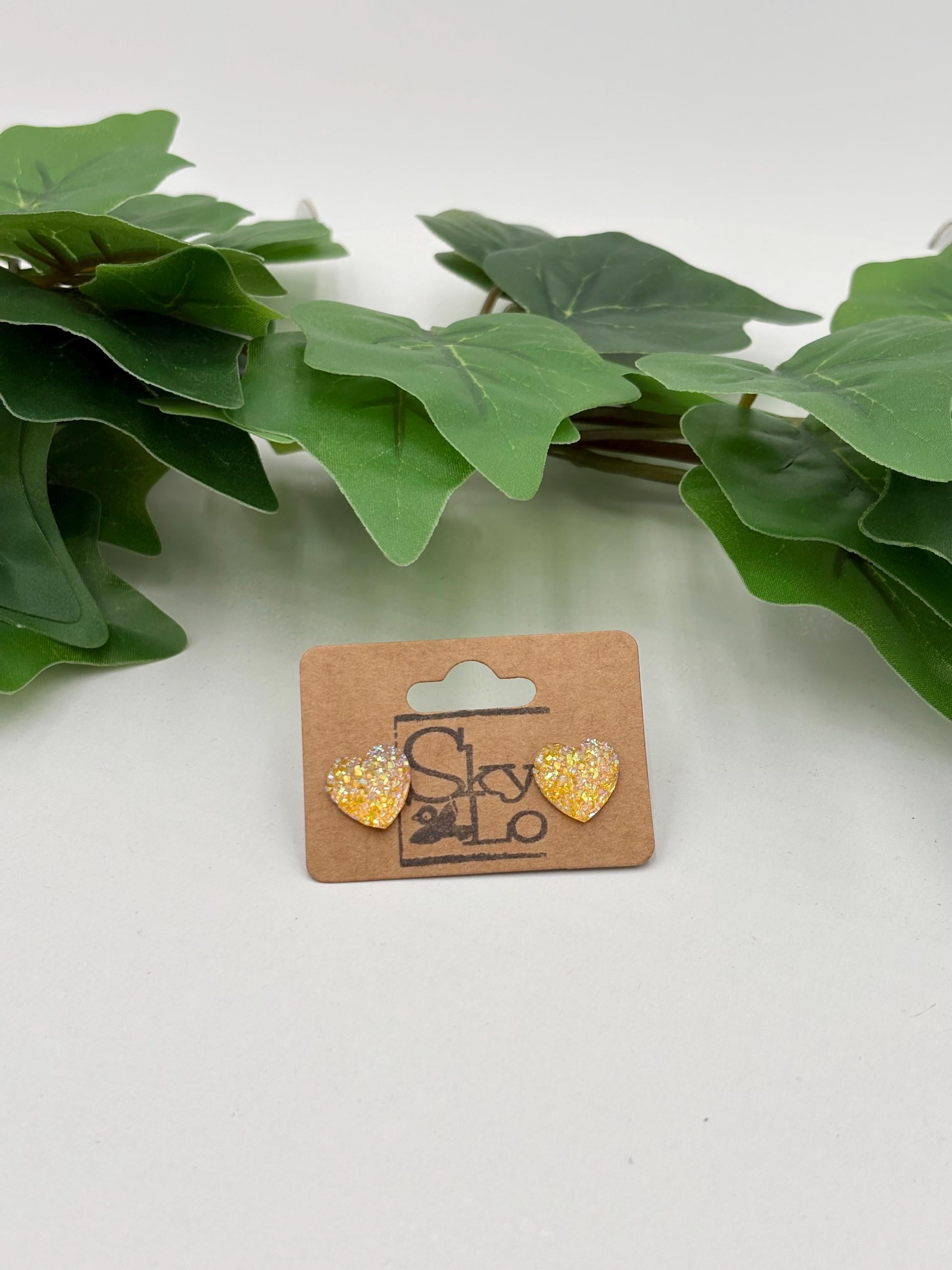 Yellow sparkling heart-shaped earrings on a 'SkyLo' branded card with green leaves in the background