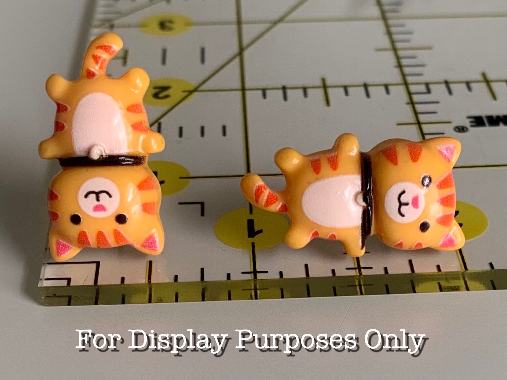 Two small orange cat earrings on a ruler with measurement markings with text "for display purposes only"