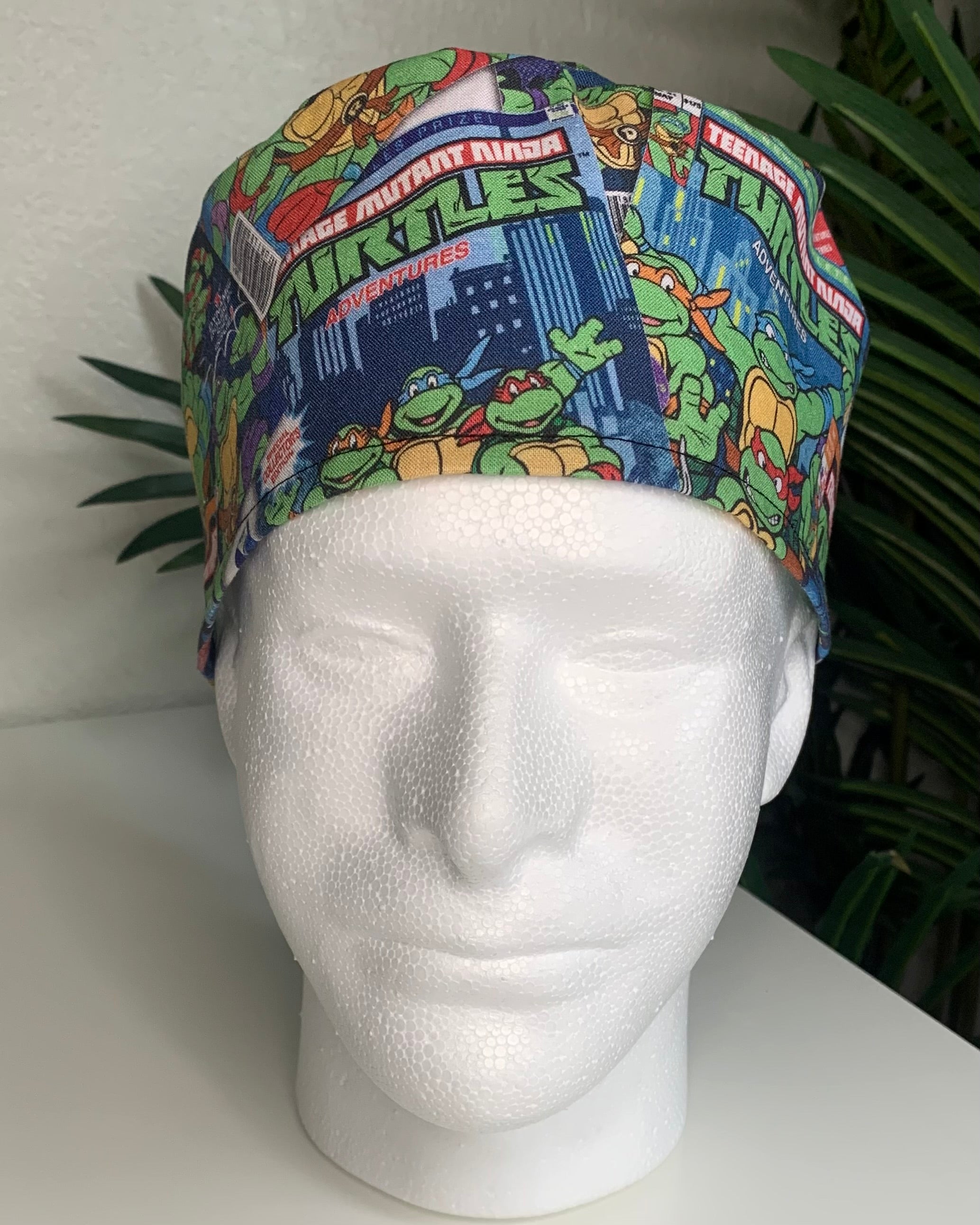 Colorful Teenage Mutant Ninja’ Turtles scrub cap on a mannequin head with a plant, against a white background