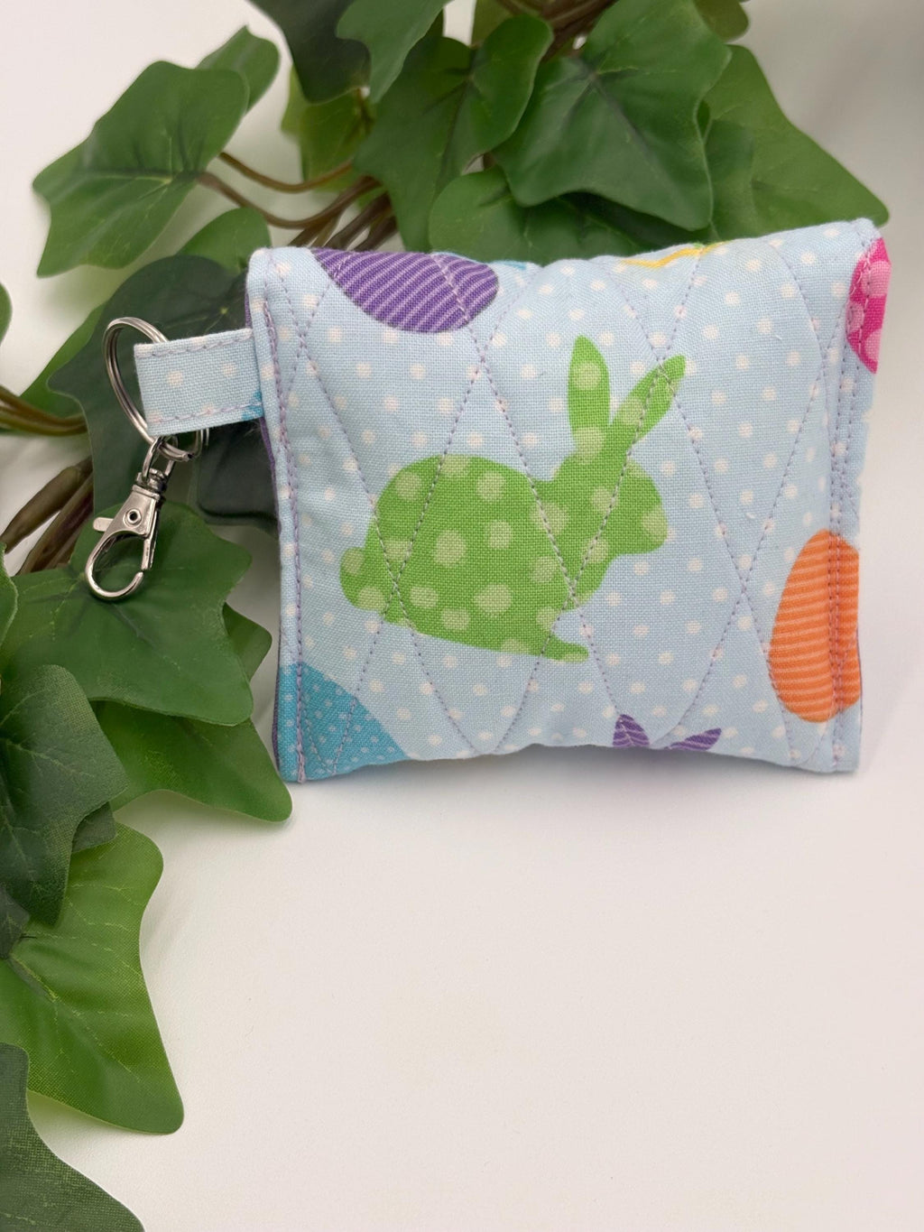 Back of a Small quilted headphone holder pouch keychain with colorful easter design on a white surface with green leaves.