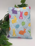Quilted book sleeve  with pastel colorful bunny and polka dot pattern on a light blue background fabric with a plant in the back and white background. 