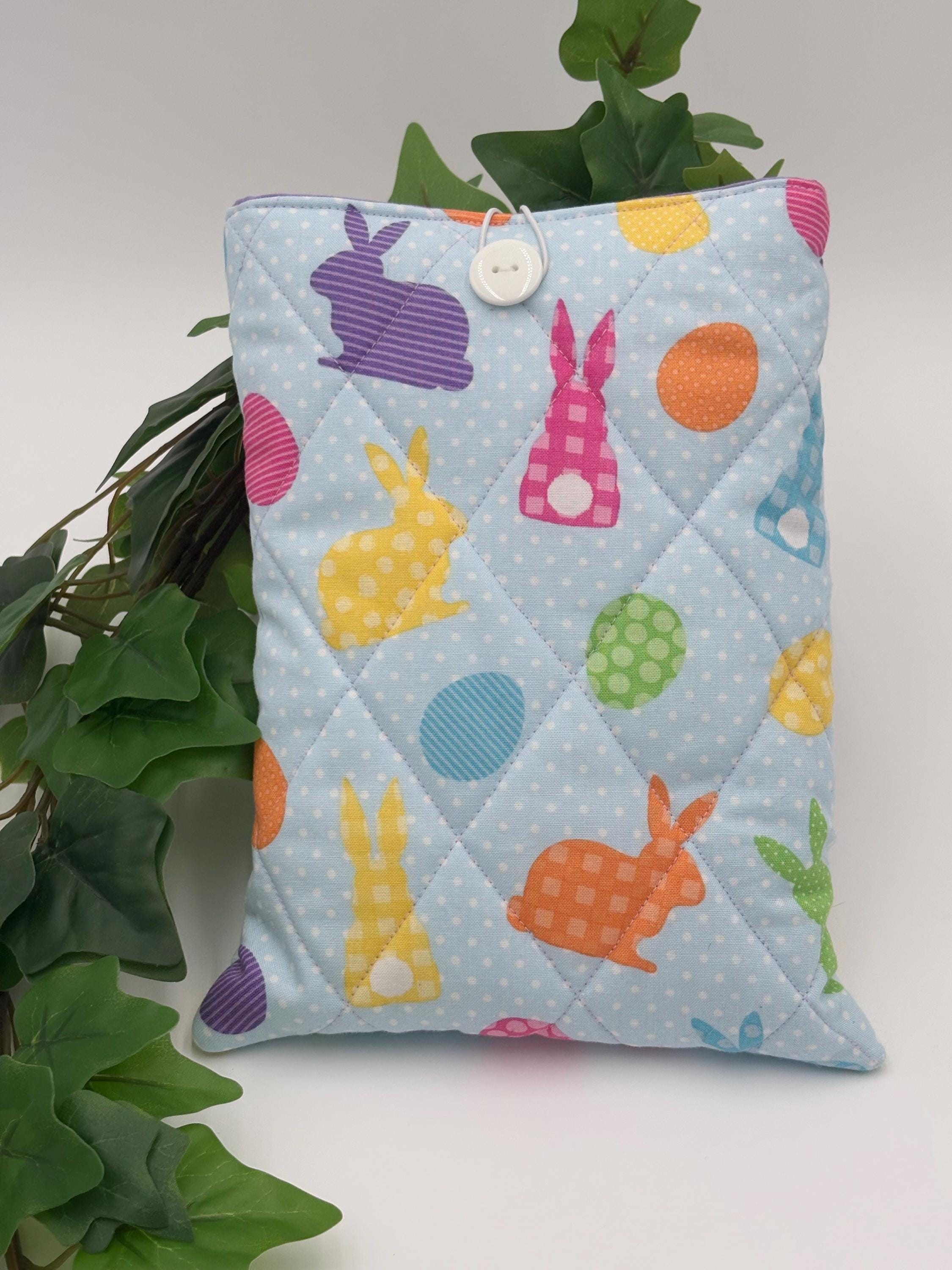 Quilted book sleeve  with pastel colorful bunny and polka dot pattern on a light blue background fabric with a plant in the back and white background. 