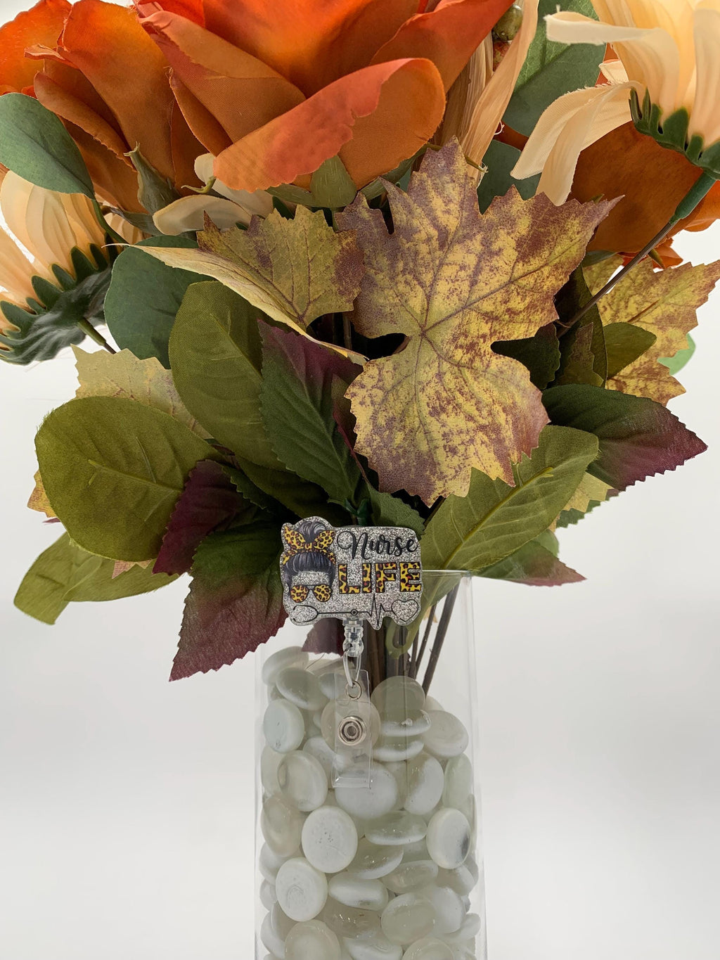 Bouquet of autumn-themed flowers in a clear vase with decorative stones on a white background with a badge reel that says Nurse Life