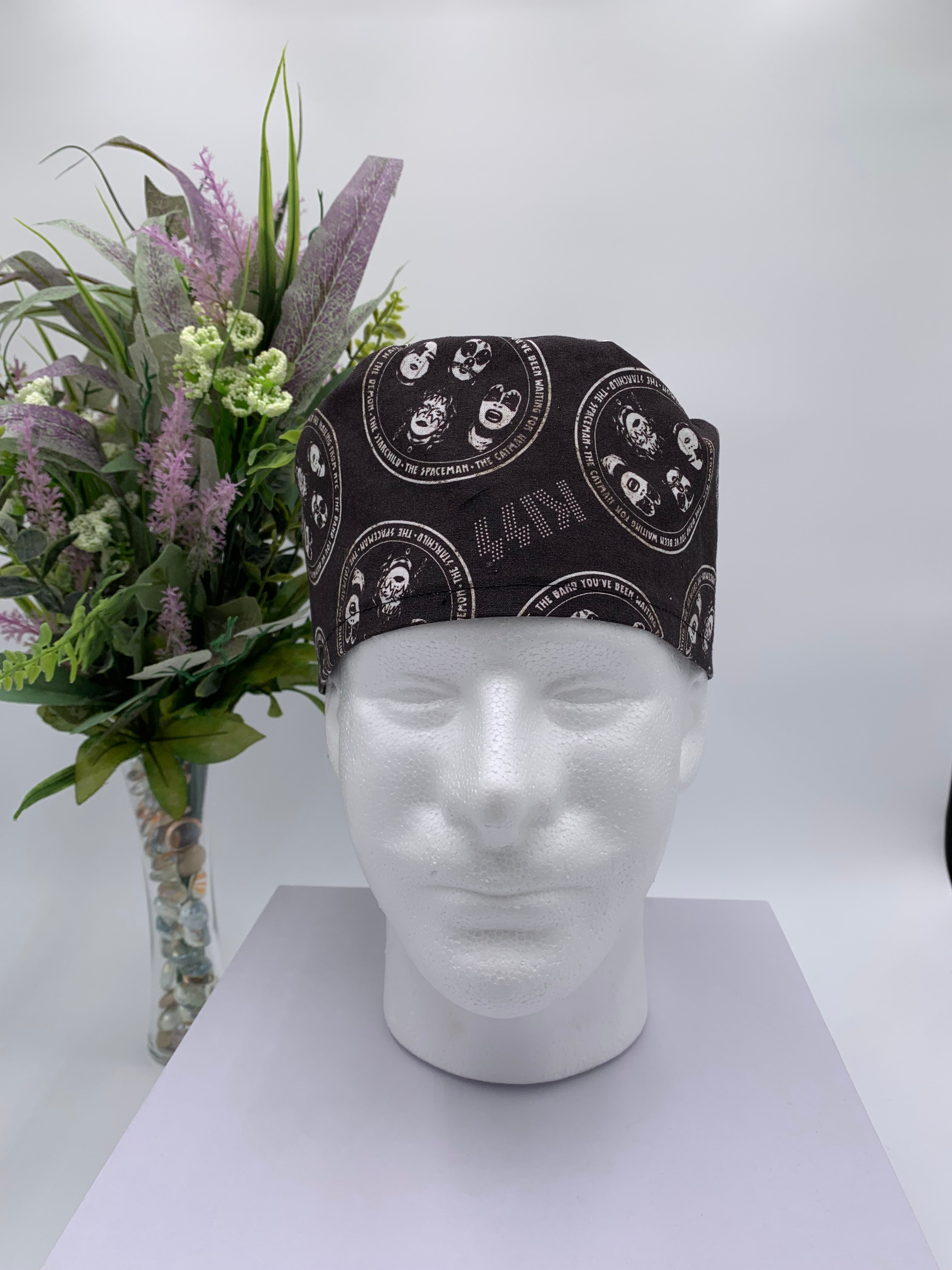Black KISS band patterned scrub cap on a mannequin head with a white background with flowers