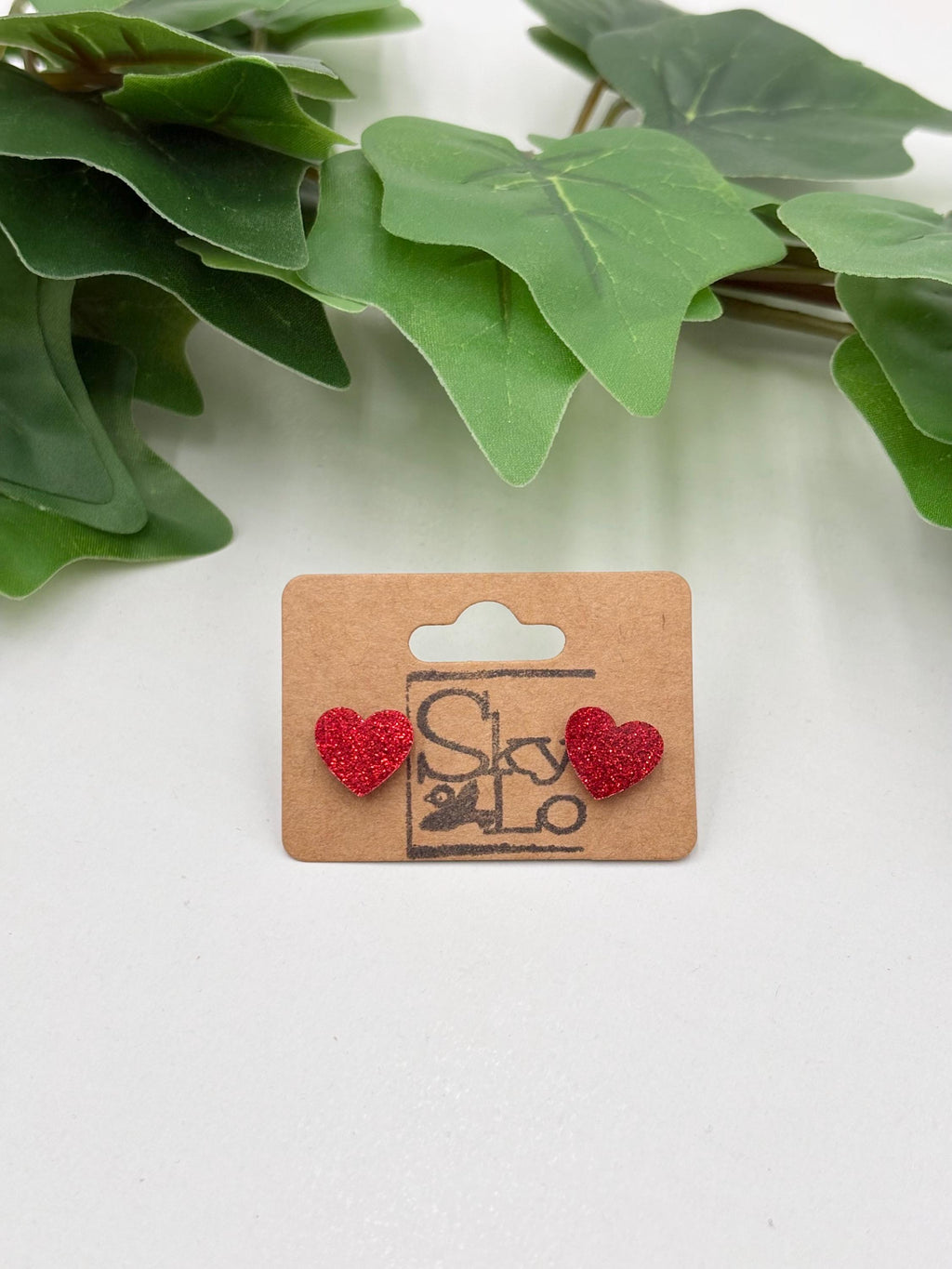 Red sparkling heart-shaped earrings on a wooden card with a brand logo, placed against green leaves.