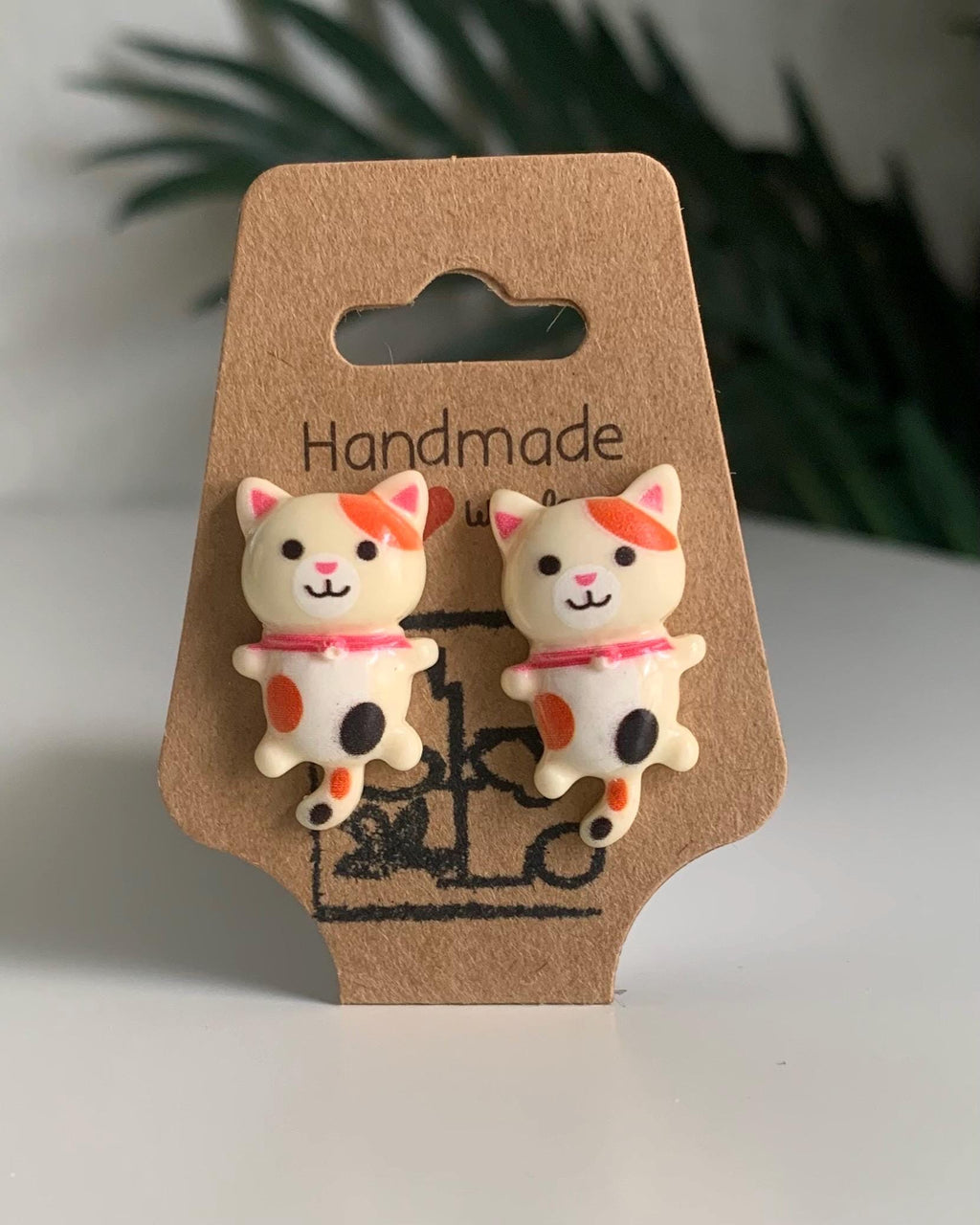 White Calico Cat-shaped earrings on a handmade cardboard backing with a blurred green plant in the background.