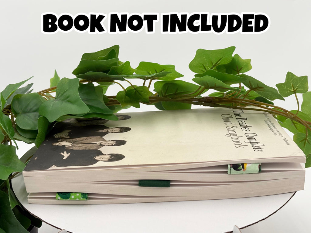 A Book with a green leafy branch on a white surface, text 'BOOK NOT INCLUDED' at the top with three green magnetic bookmarks on pages. 