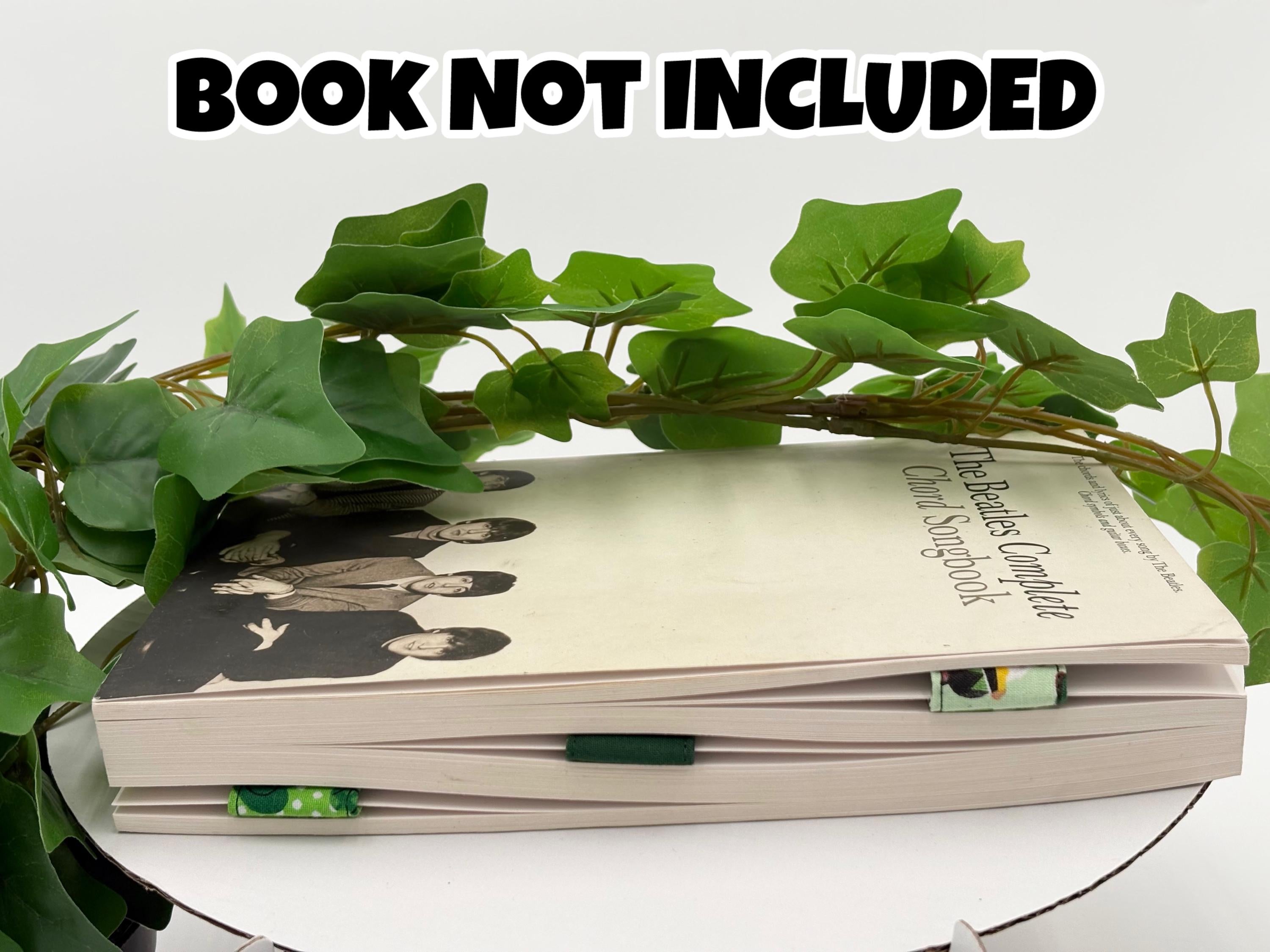 A Book with a green leafy branch on a white surface, text 'BOOK NOT INCLUDED' at the top with three green magnetic bookmarks on pages. 
