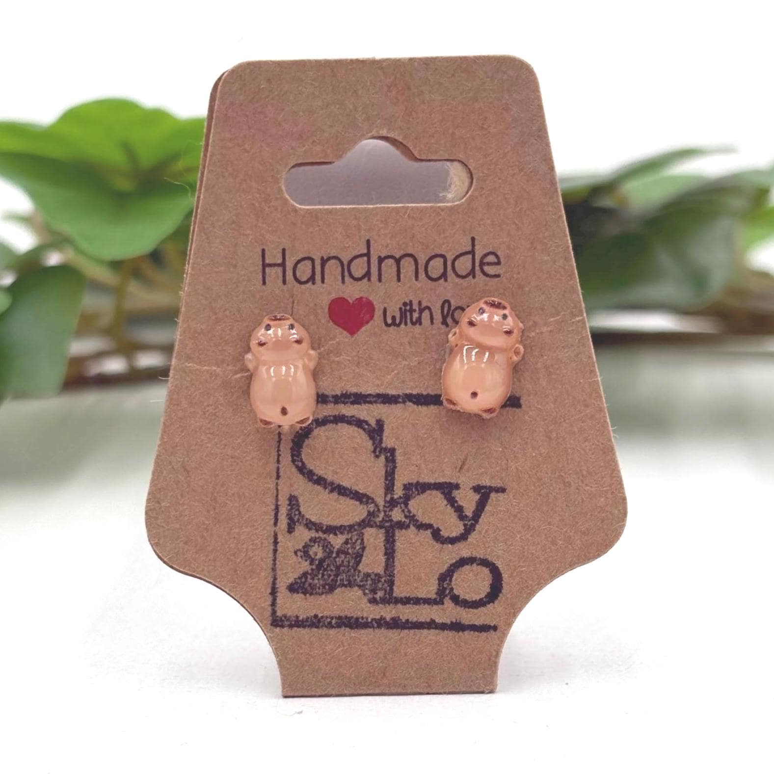Handmade capybara laying down earrings on a branded card with a blurred green plant background