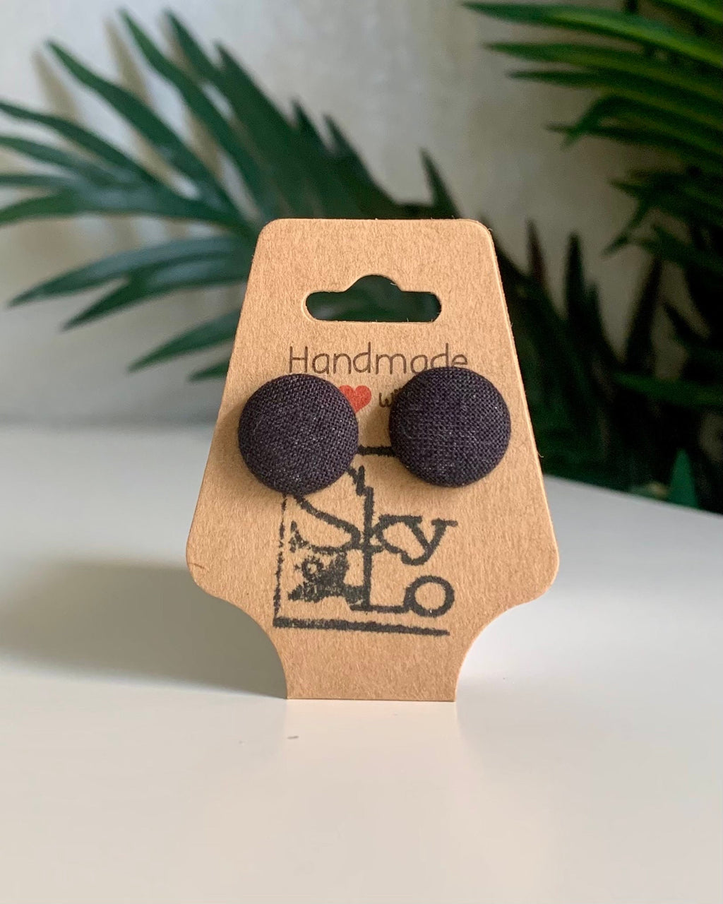 Pair of black round earrings on a 'Handmade' branded card with a plant in the background.