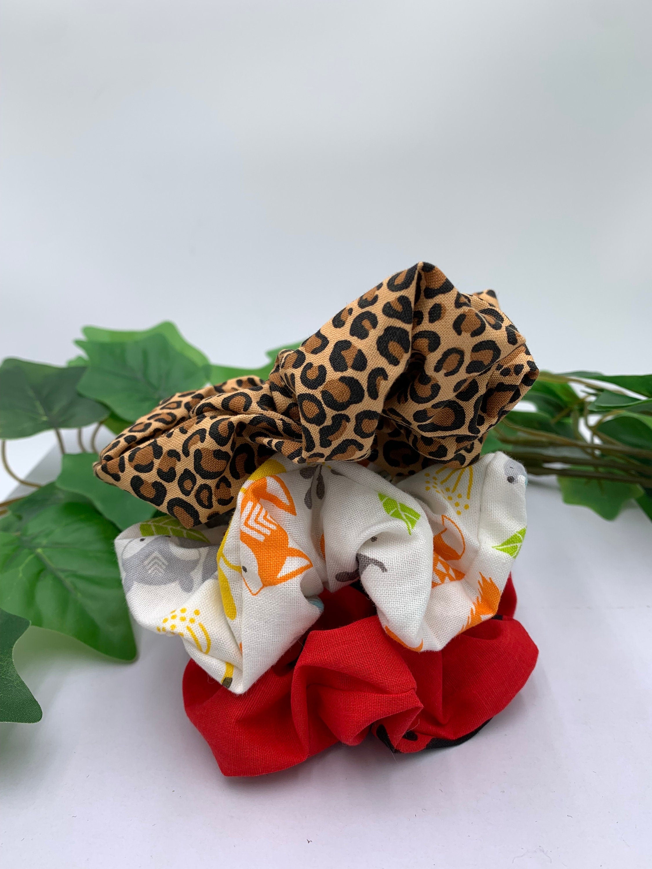 Set of three scrunchies with leopard print, white patterned, and red designs on a white background next to green leaves
