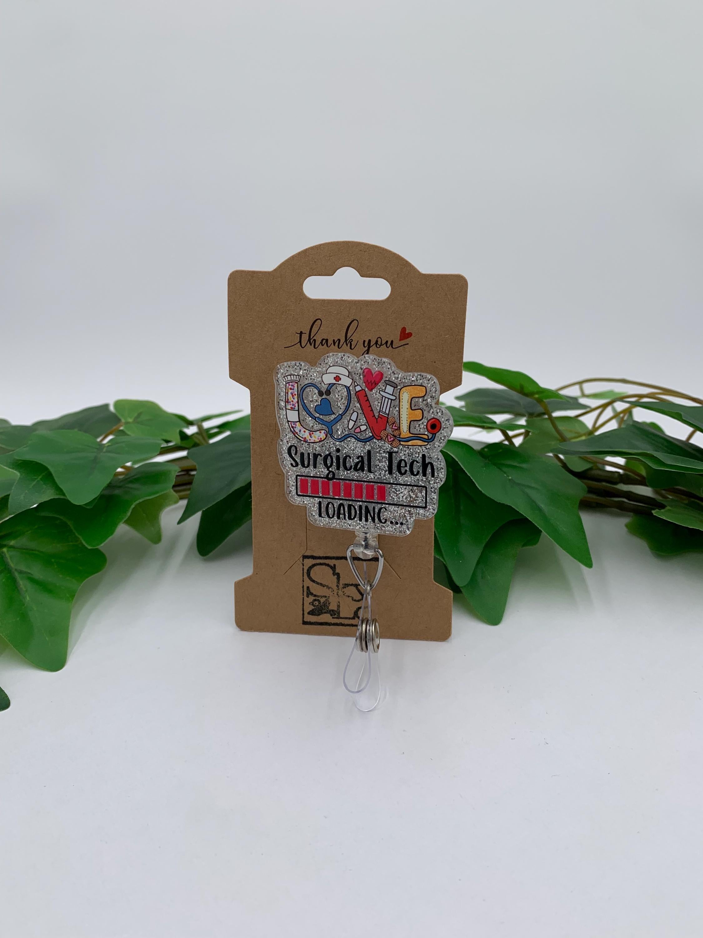 Badge reel with 'Surgical Tech Loading' design on a cardboard backing, surrounded by green leaves.