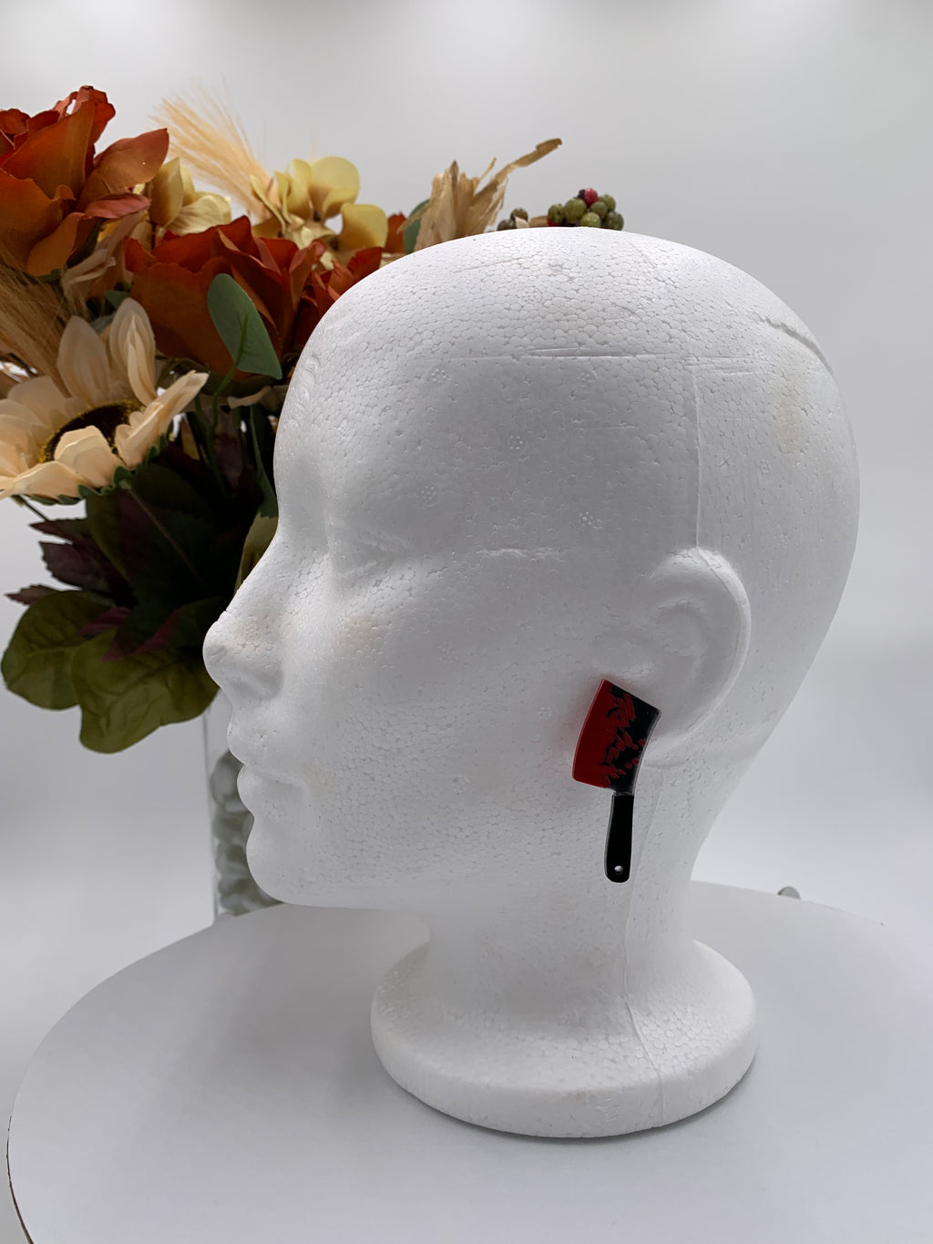 White Mannequin head with bloody meat cleaver earrings on a white background with a bouquet of flowers. 