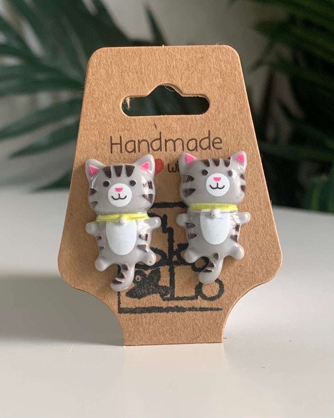Gray American Short Hair Cat-shaped earrings on a 'Handmade' branded card with a blurred green plant background.