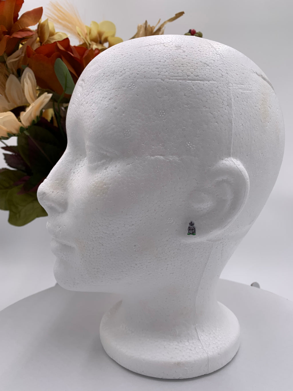 White mannequin head with a small tombstone earring on a white background with a bouquet of flowers