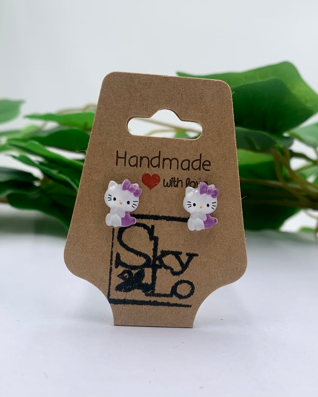 Handmade Hello Kitty earrings on a brown card with green leaves in the background