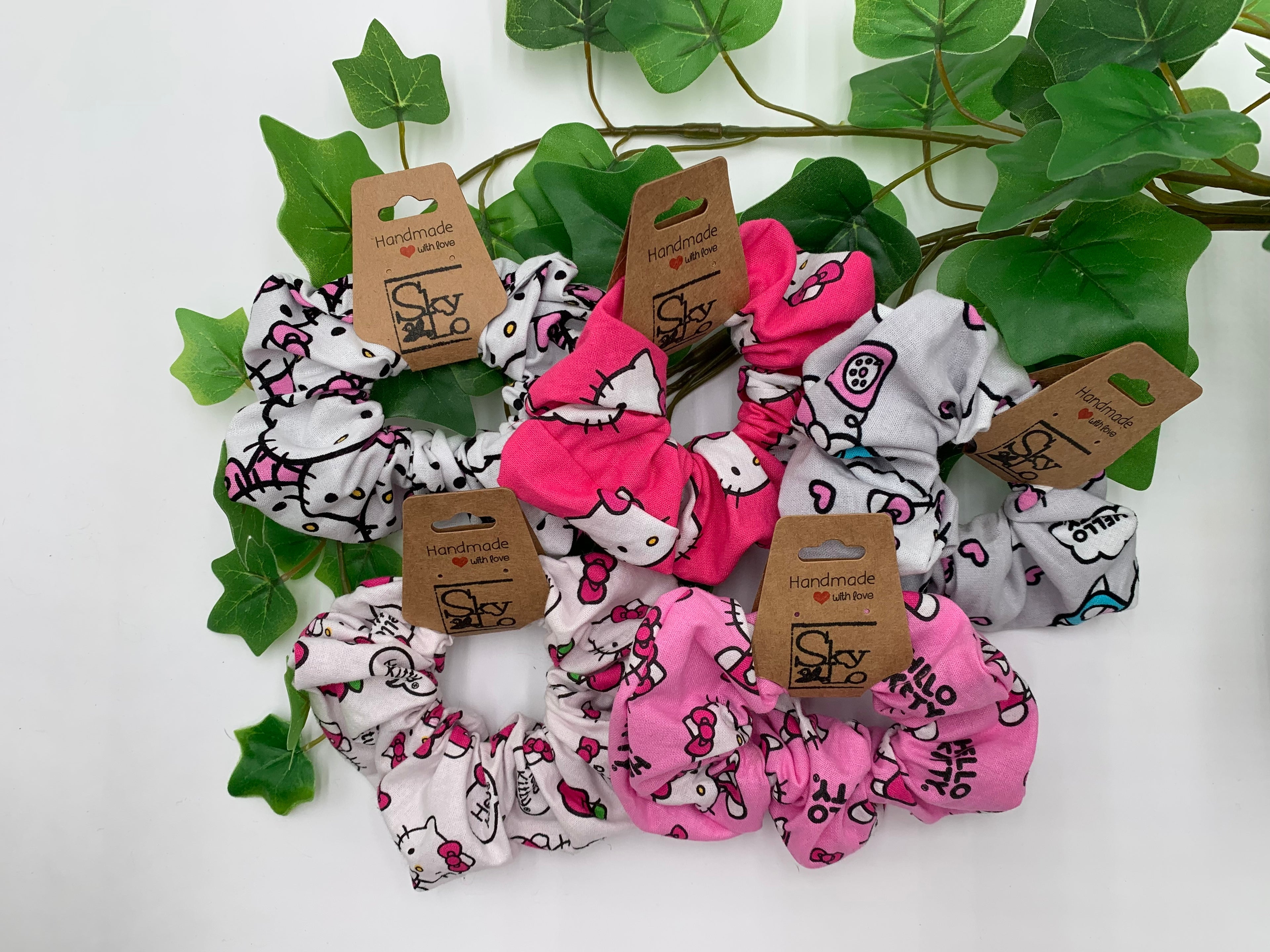 Set of 5 scrunchies with cute cat designs on a white background with green leaves.