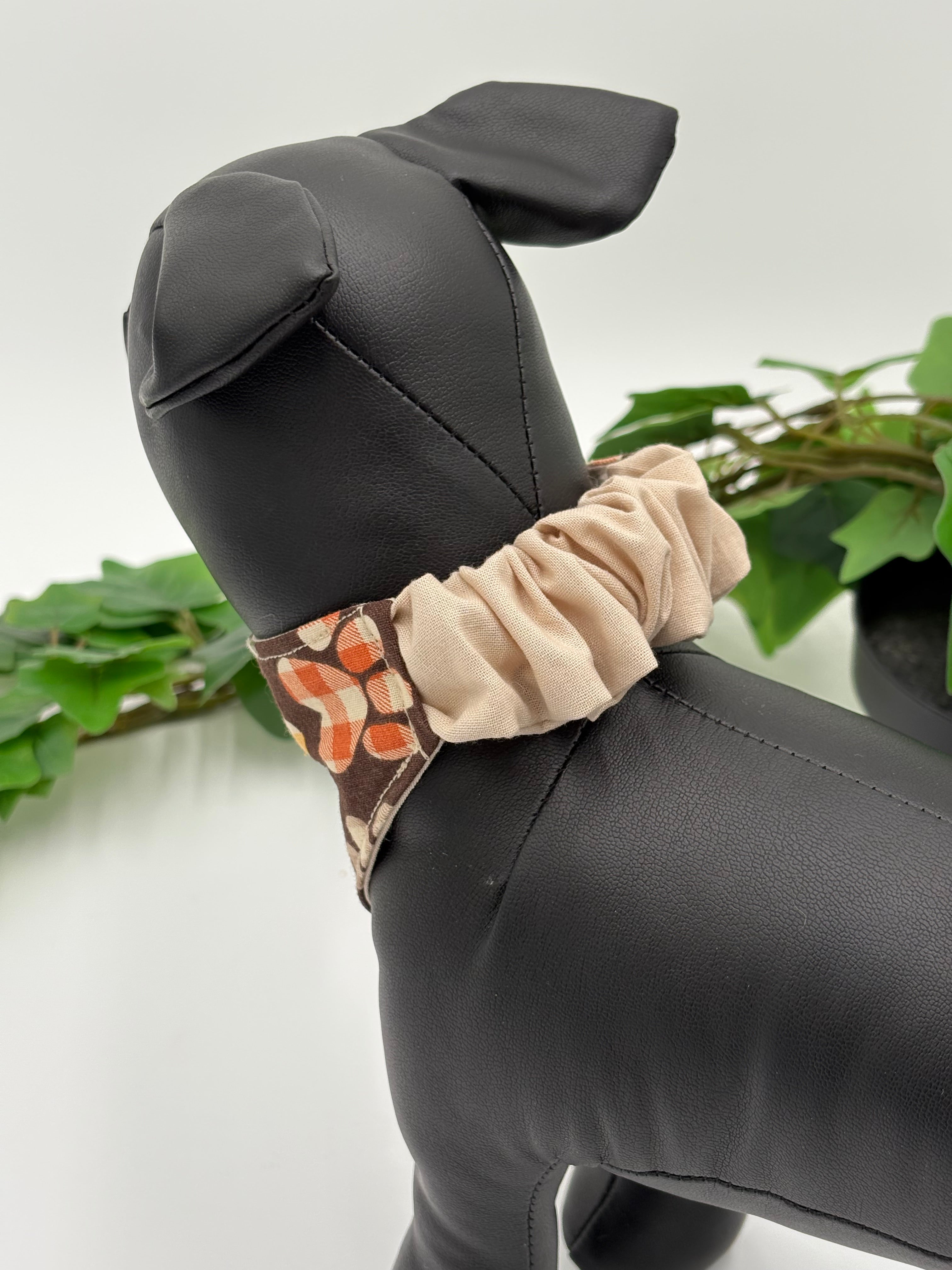 Black dog mannequin wearing a brown with plaid paw prints bandana with a khaki colored scrunchie on a white background with green leaves.
