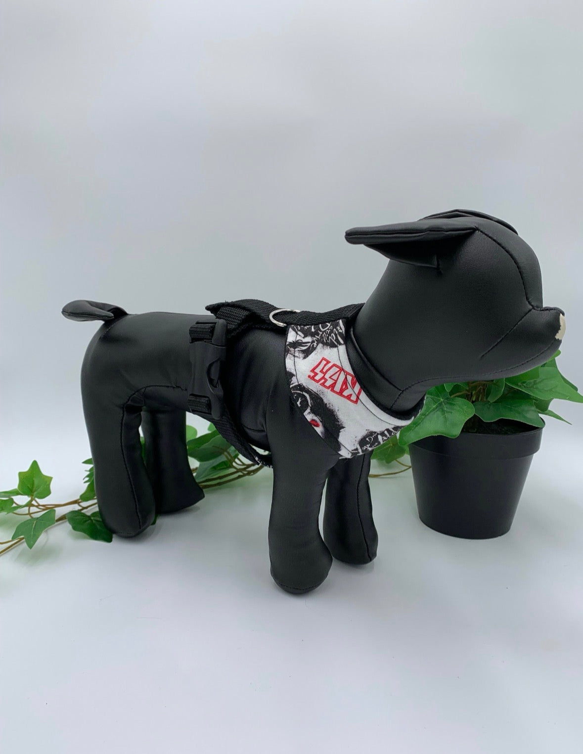 Black dog mannequin with white heavy metal band pattern harness on a white background with a potted green plant