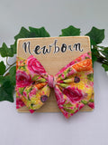 Floral big bow headband on a wooden board with 'Newborn' text, surrounded by green leaves.