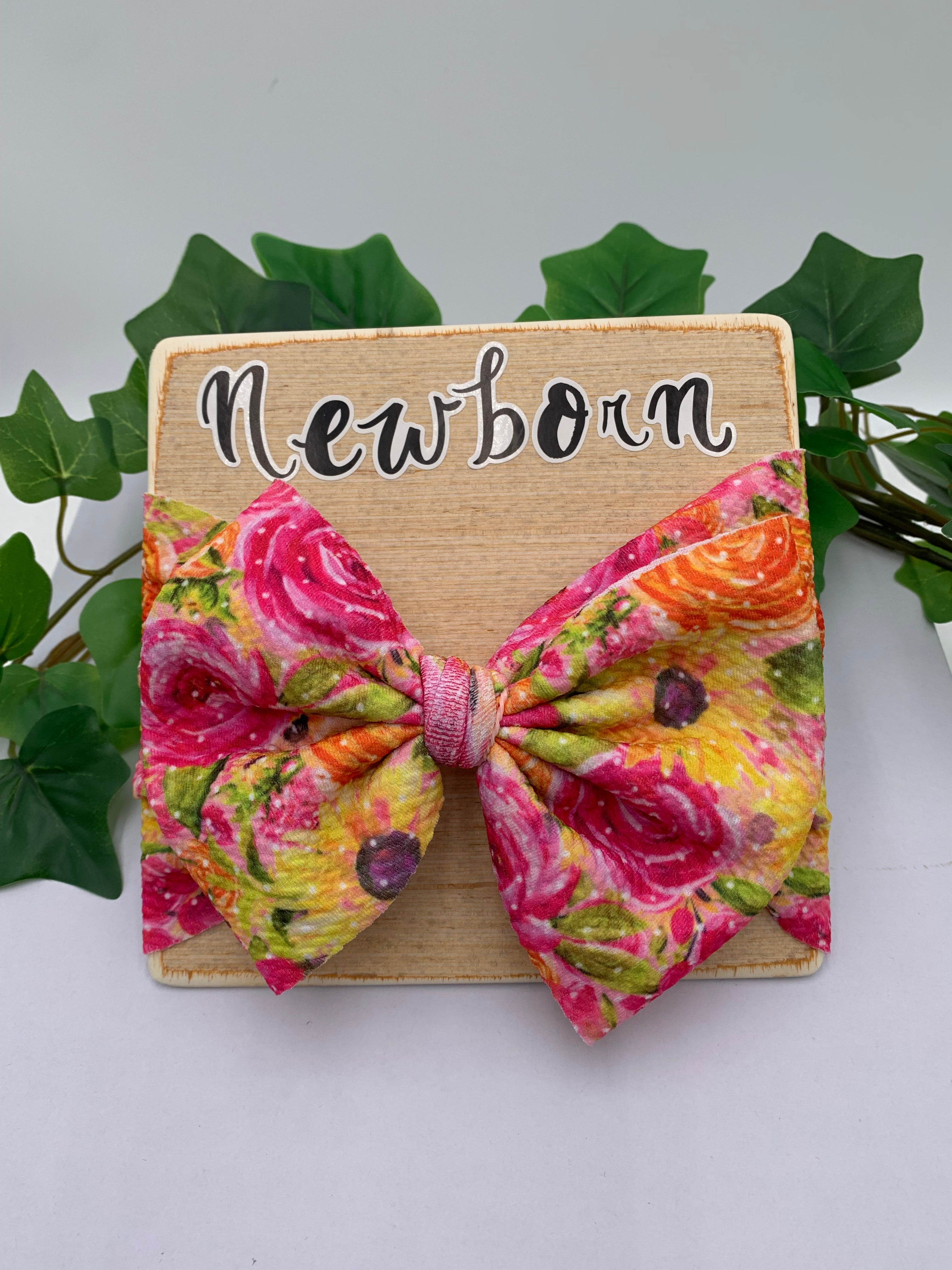 Floral big bow headband on a wooden board with 'Newborn' text, surrounded by green leaves.