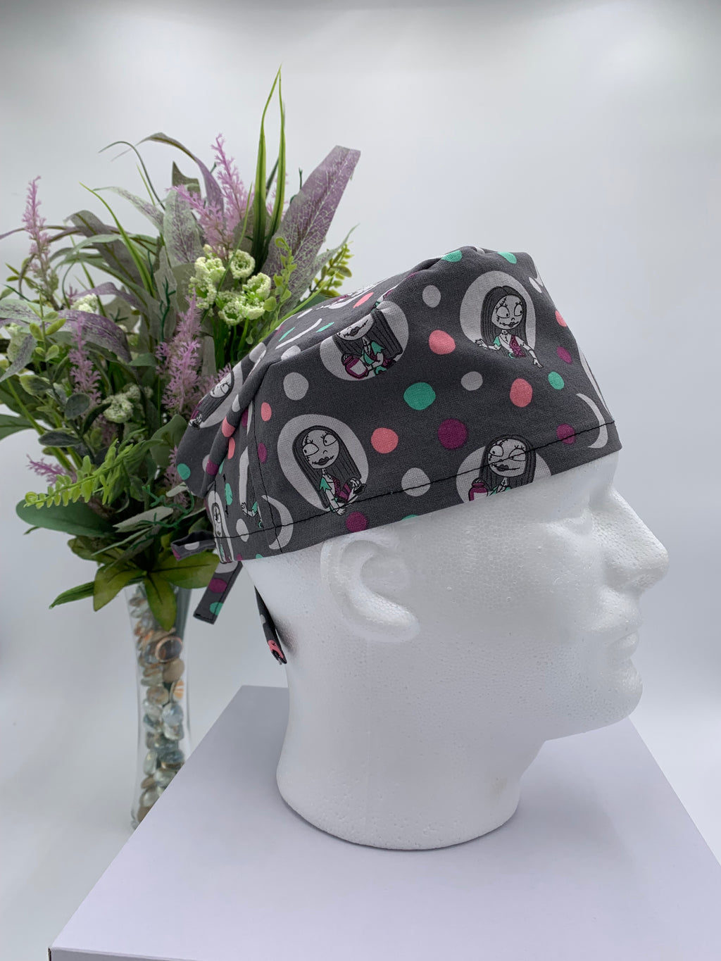 Gray polka dot nightmare before Christmas scrub cap on a mannequin head with flowers, against a white background