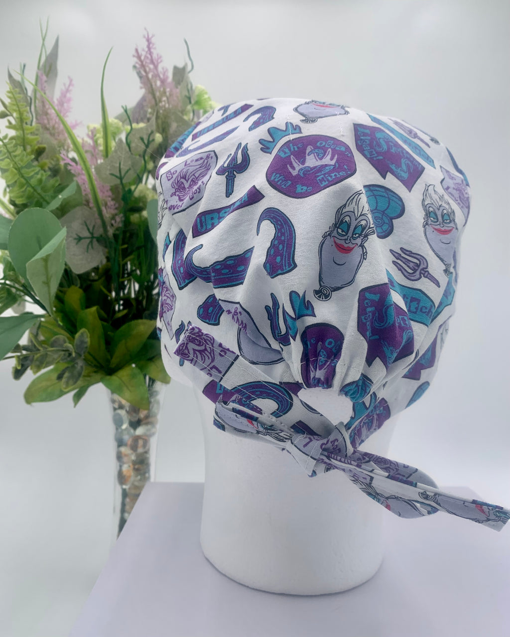 Colorful Little Mermaid Ursula scrub cap on a mannequin head with flowers, against a white background