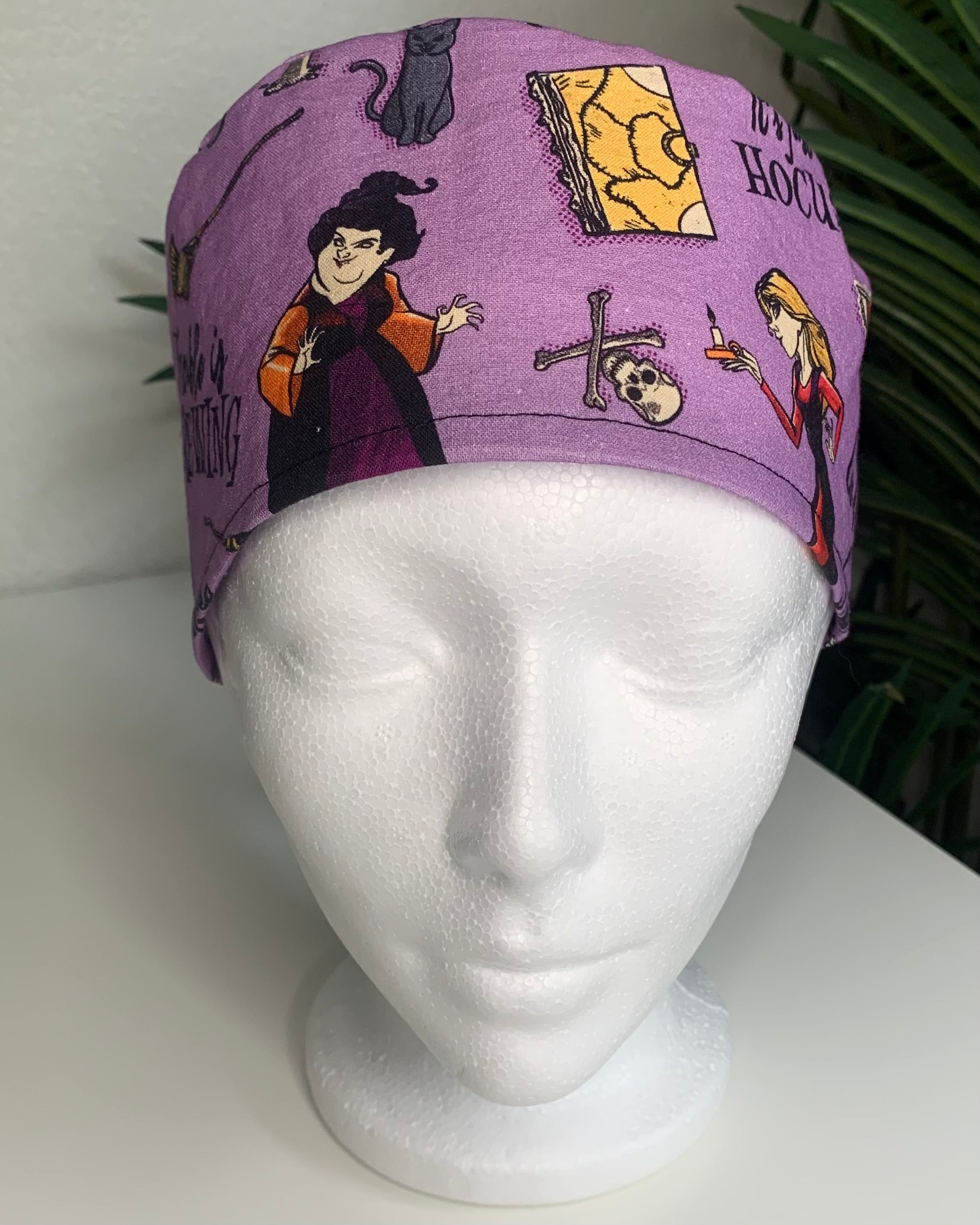 Purple Hocus Pocus patterned scrub cap on a mannequin head with a white background with plant