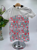 White t-shirt with a colorful valentines gnome and heart pattern on against a white background with a vase of flowers