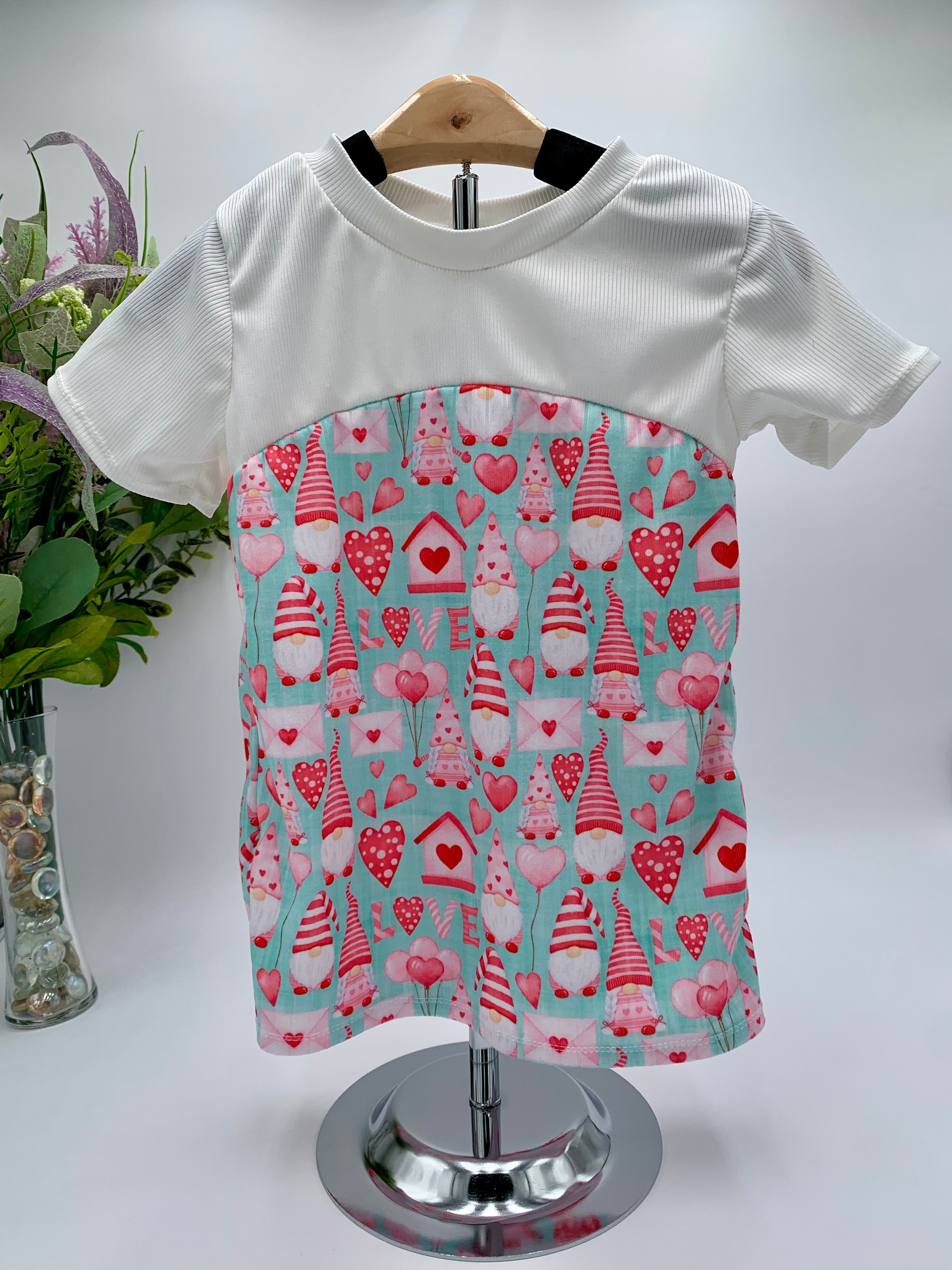 White t-shirt with a colorful valentines gnome and heart pattern on against a white background with a vase of flowers