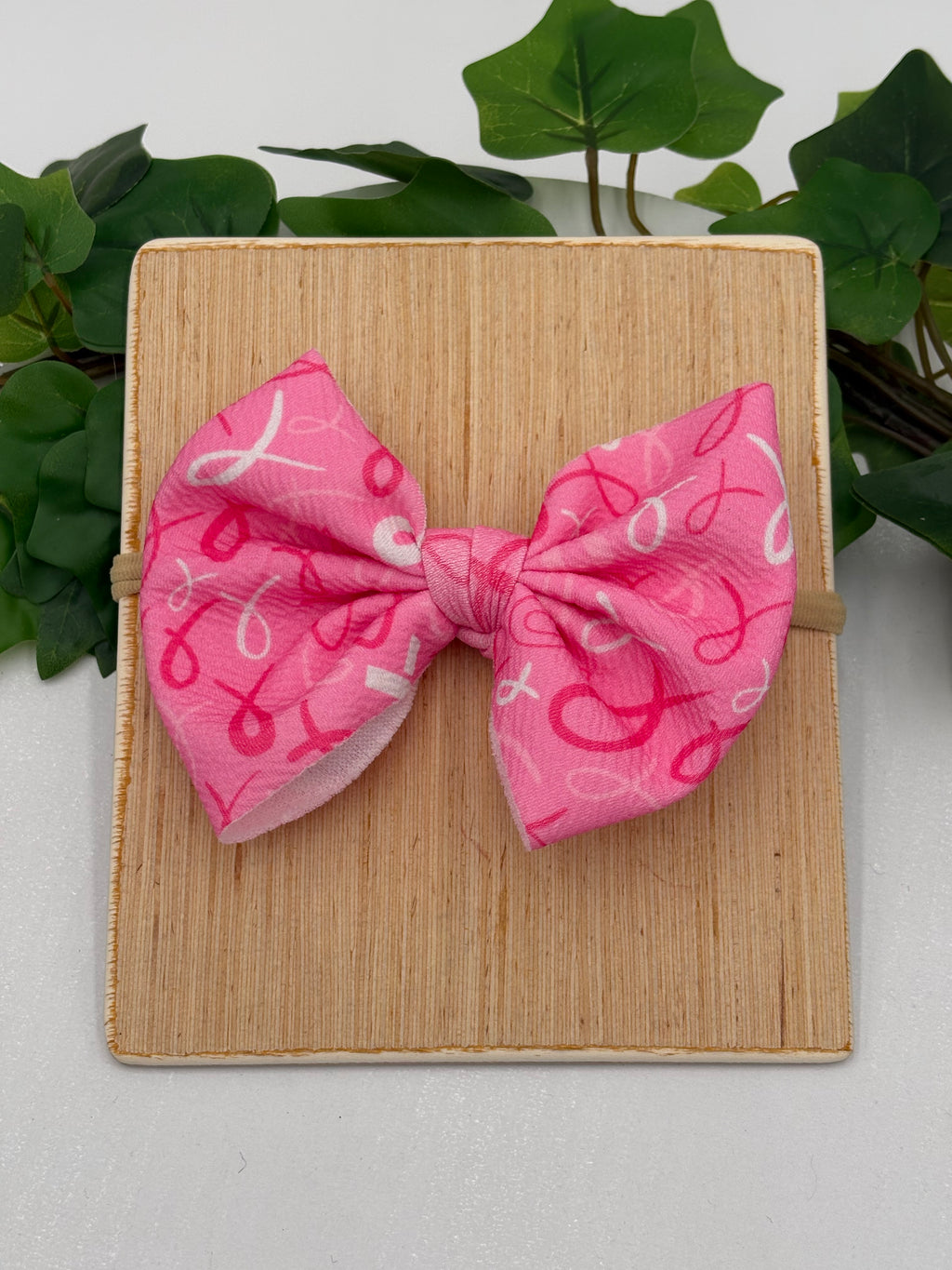 Pink Breast Cancer awareness themed big bow nylon headband on a wooden board with a white background and greenery 