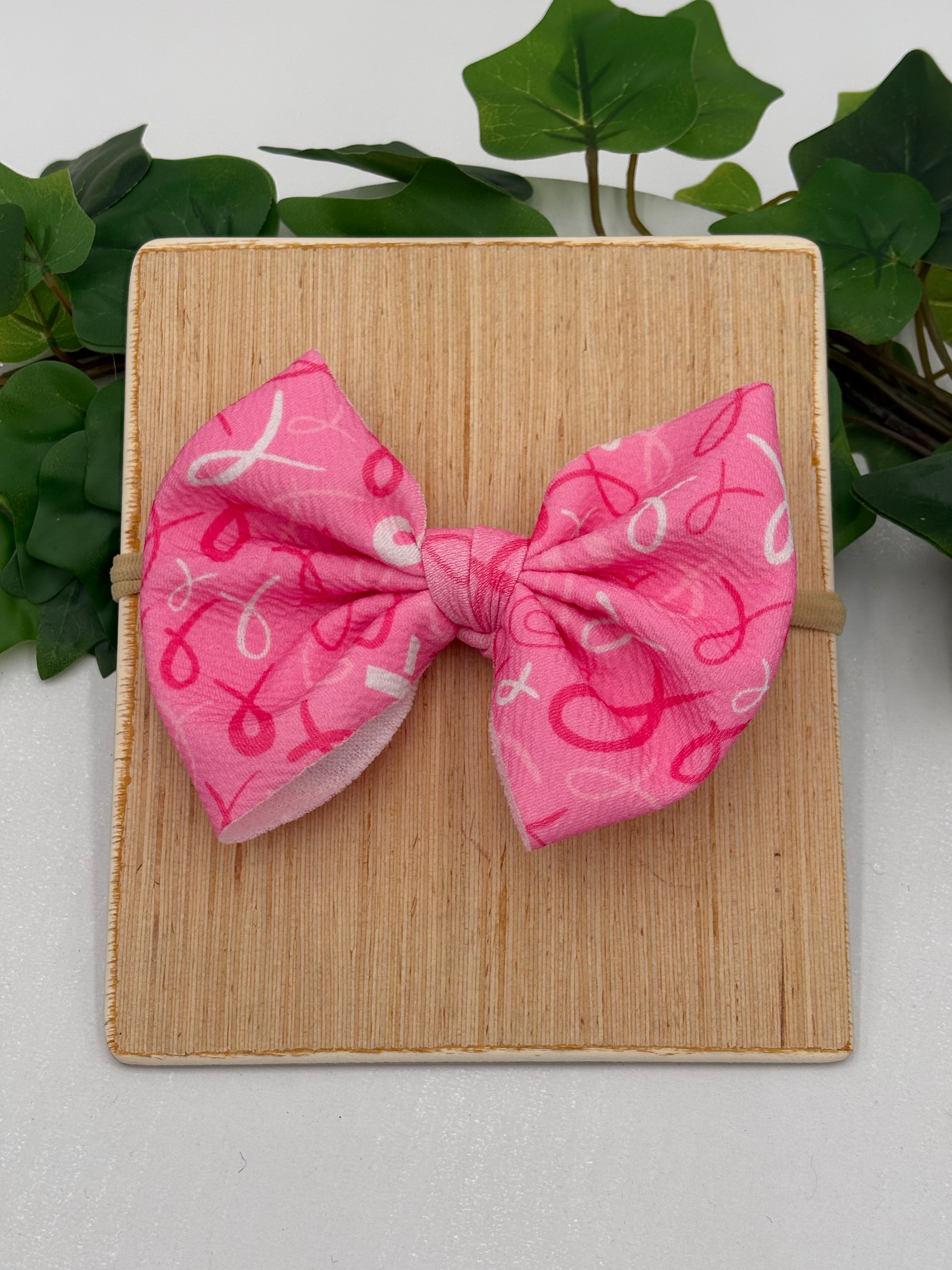 Pink Breast Cancer awareness themed big bow nylon headband on a wooden board with a white background and greenery 
