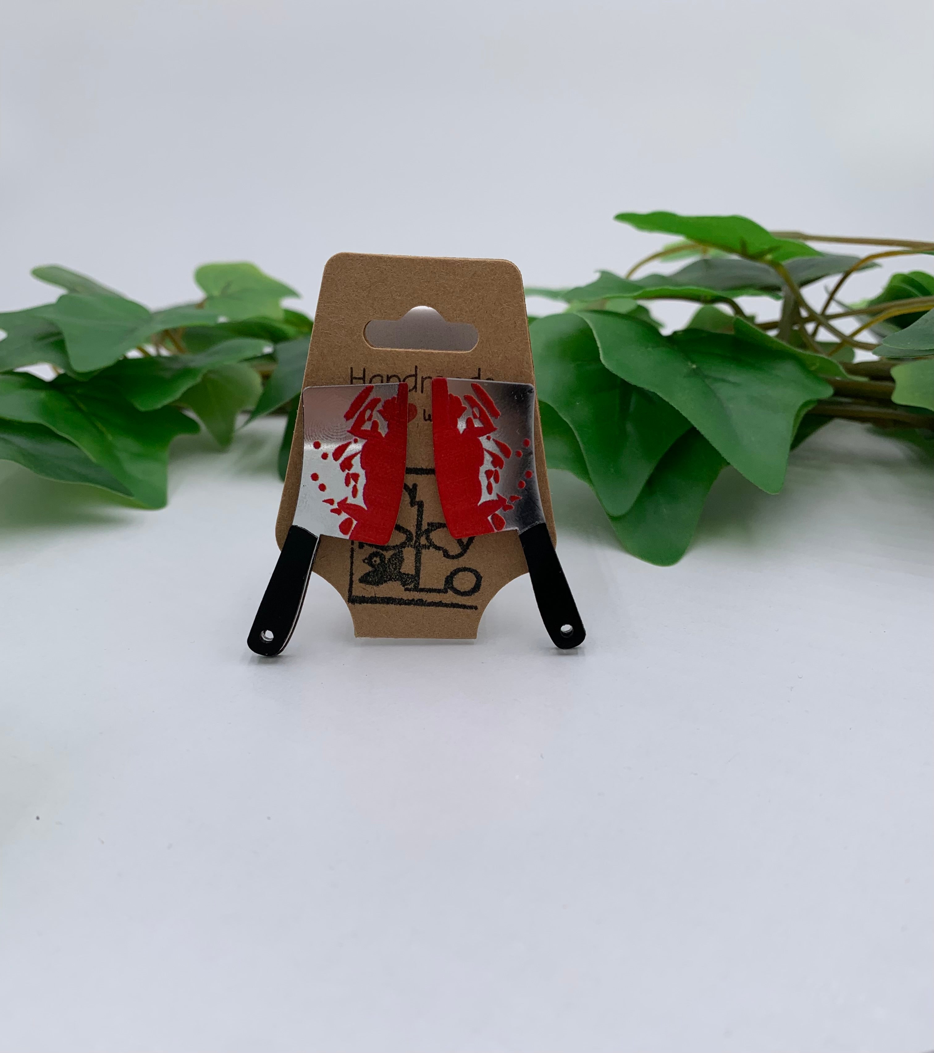 Small bloody meat cleaver knife earrings on a Kraft branded card with green leaves in the background