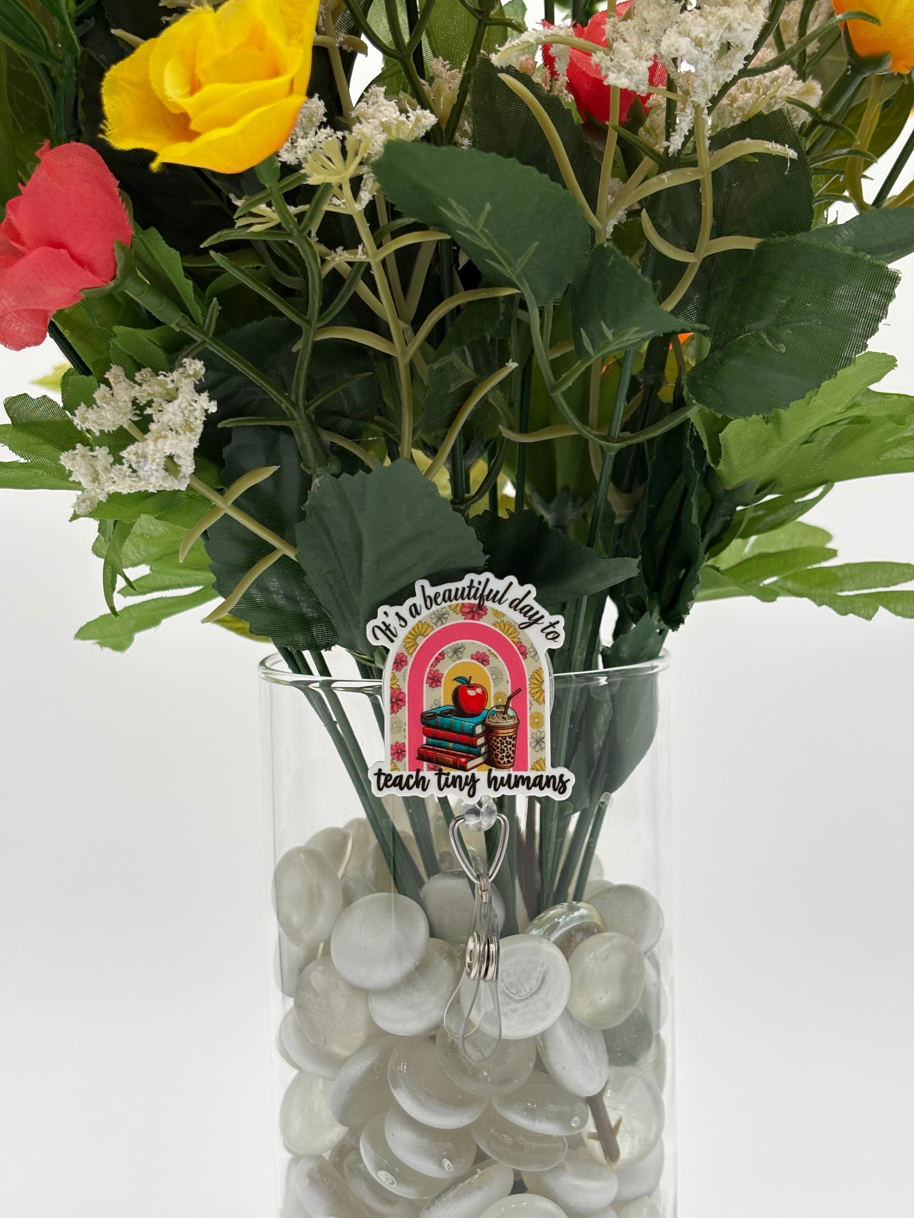 Bouquet of flowers with floral rainbow with books, an apple, and a drink with text "It's a beautiful day to teach tiny humans" badge holder in a clear vase on a white background