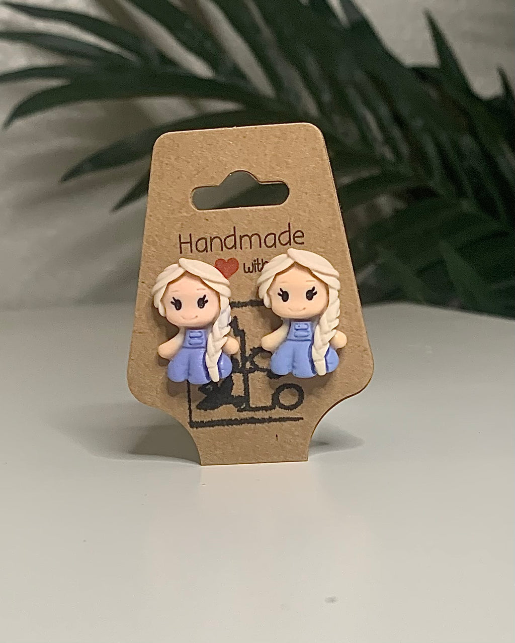 Princess shaped earrings with a Kraft branded label on a neutral background with a green plant. 