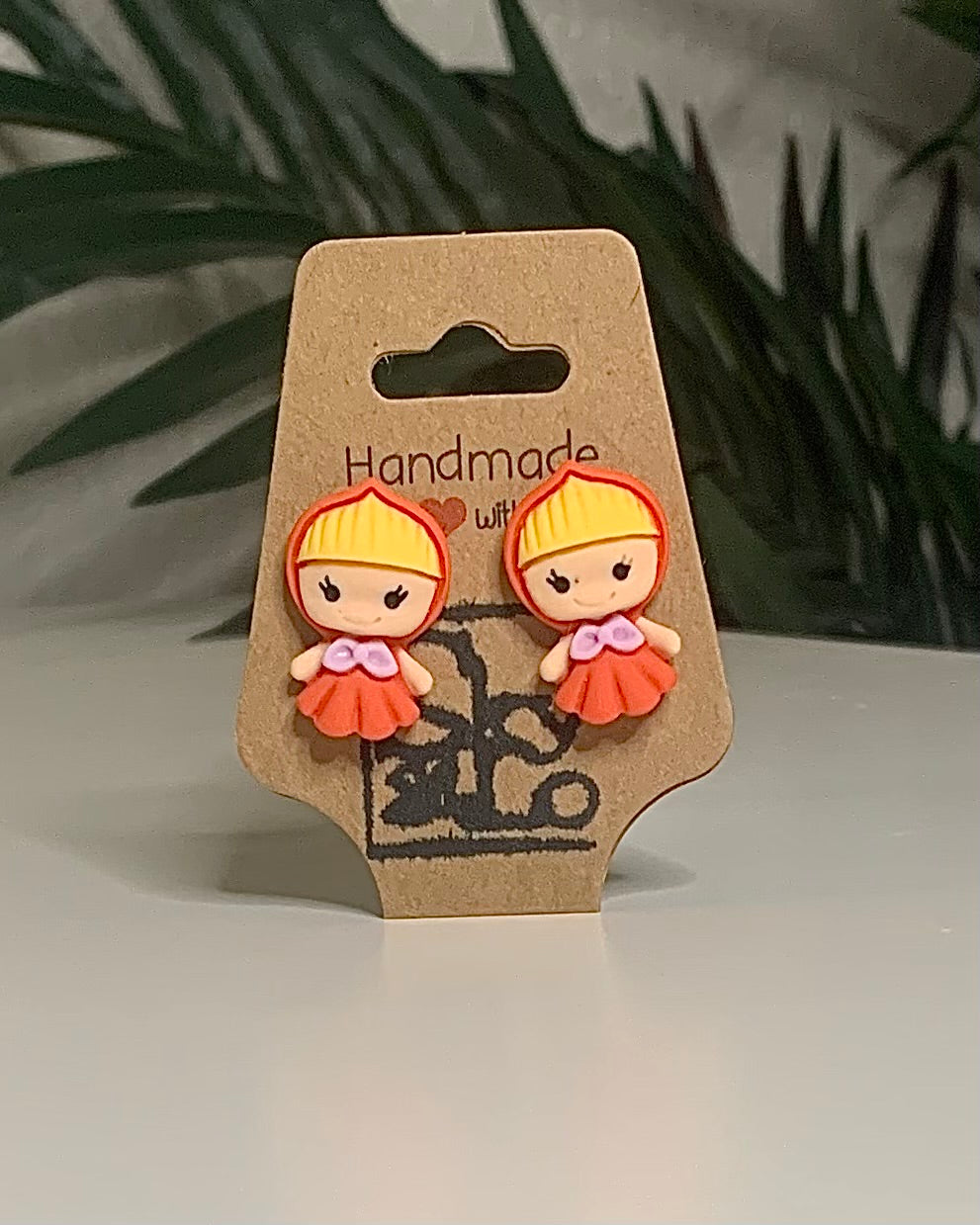 Little red girl shaped earrings with a Kraft branded label on a neutral background with a green plant. 