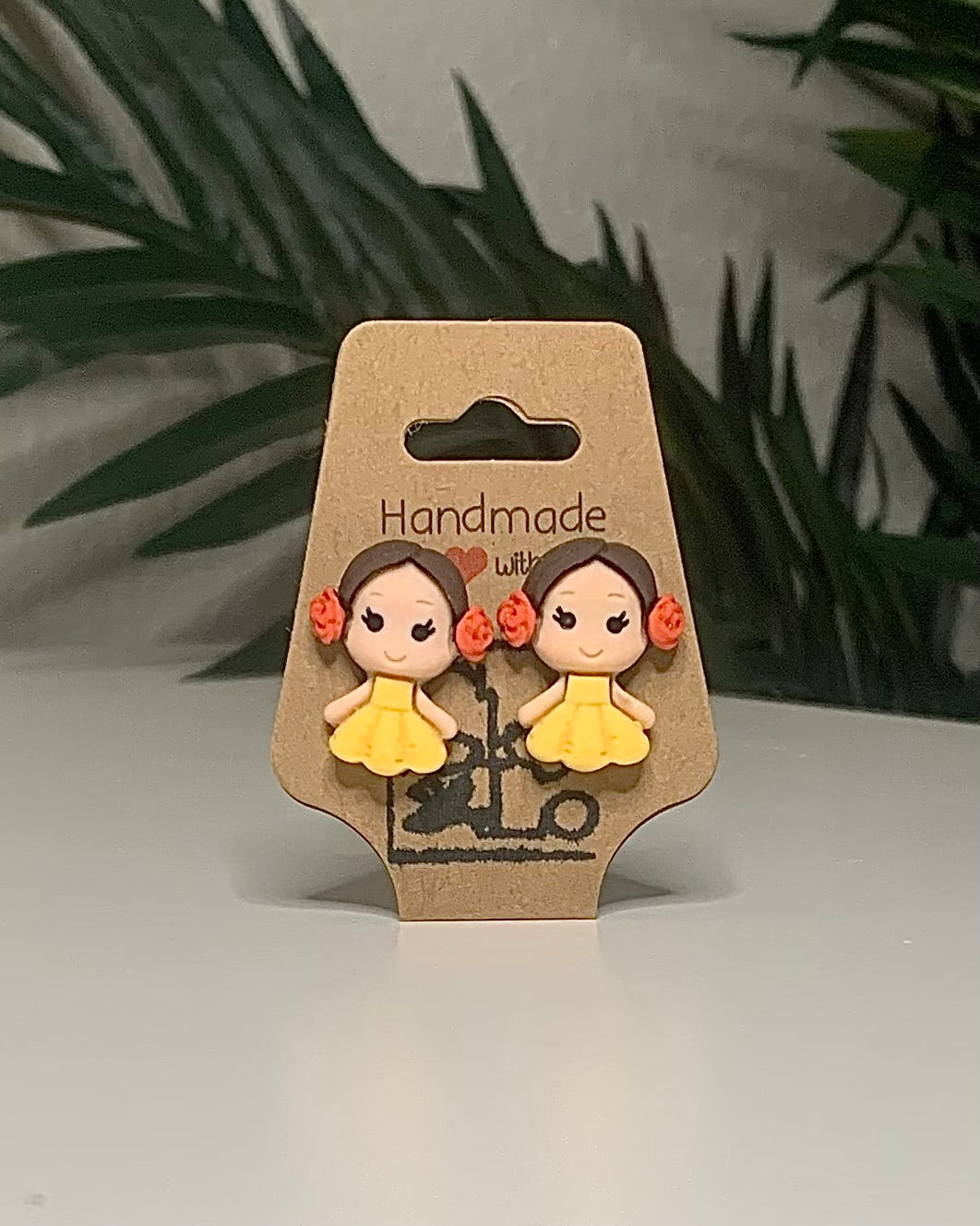 Princess shaped earrings with a Kraft branded label on a neutral background with a green plant. 