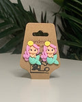 Mermaid shaped earrings with a Kraft branded label on a neutral background with a green plant. 