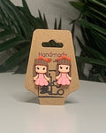 Princess shaped earrings with a Kraft branded label on a neutral background with a green plant. 