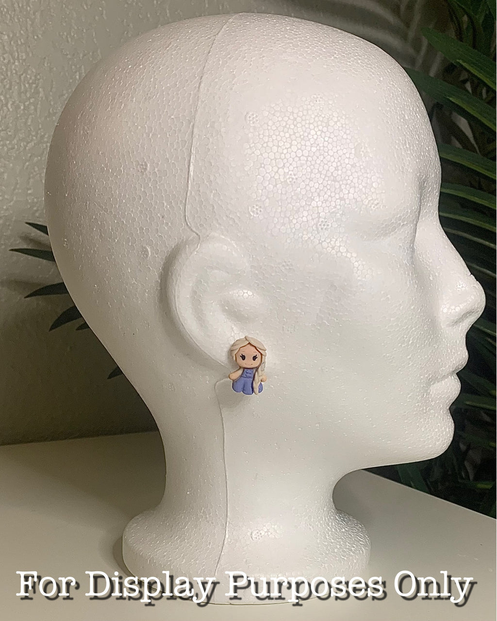 White Mannequin head with princess shaped earrings on a neutral background with a green plant. 