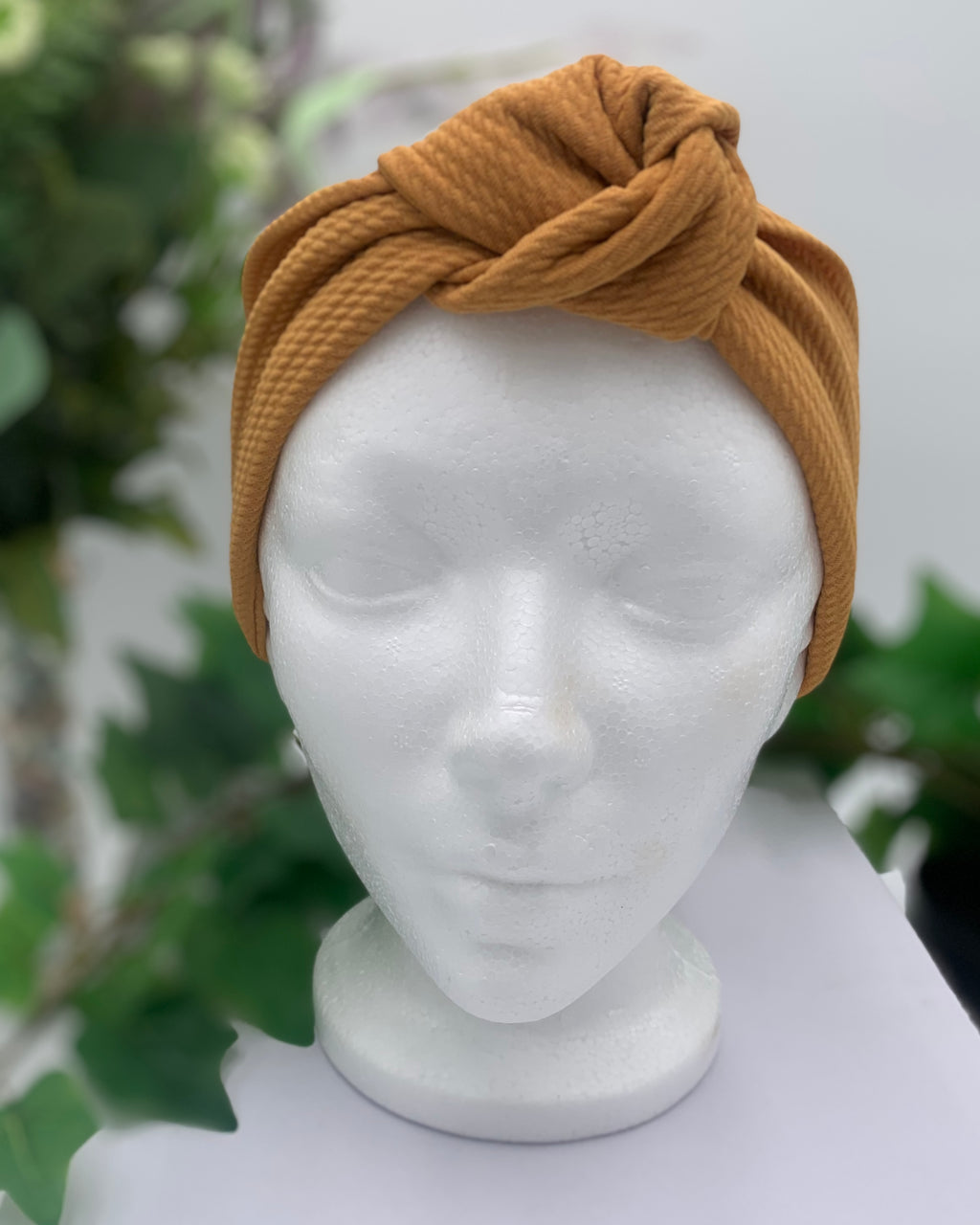 Mustard colored knot headband on a white mannequin head against a white background with green leaves