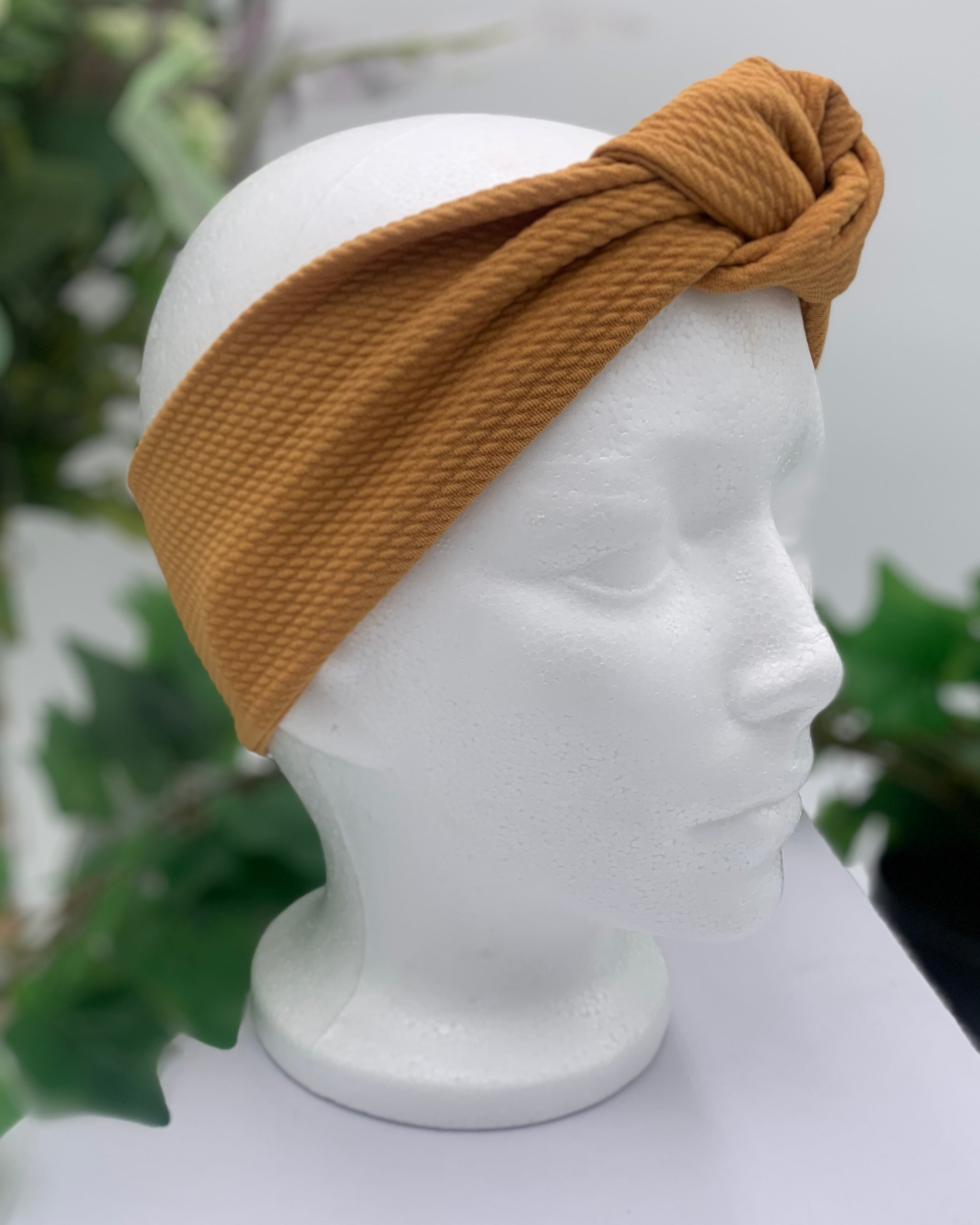 side view of a Mustard colored knot headband on a white mannequin head against a white background with green leaves