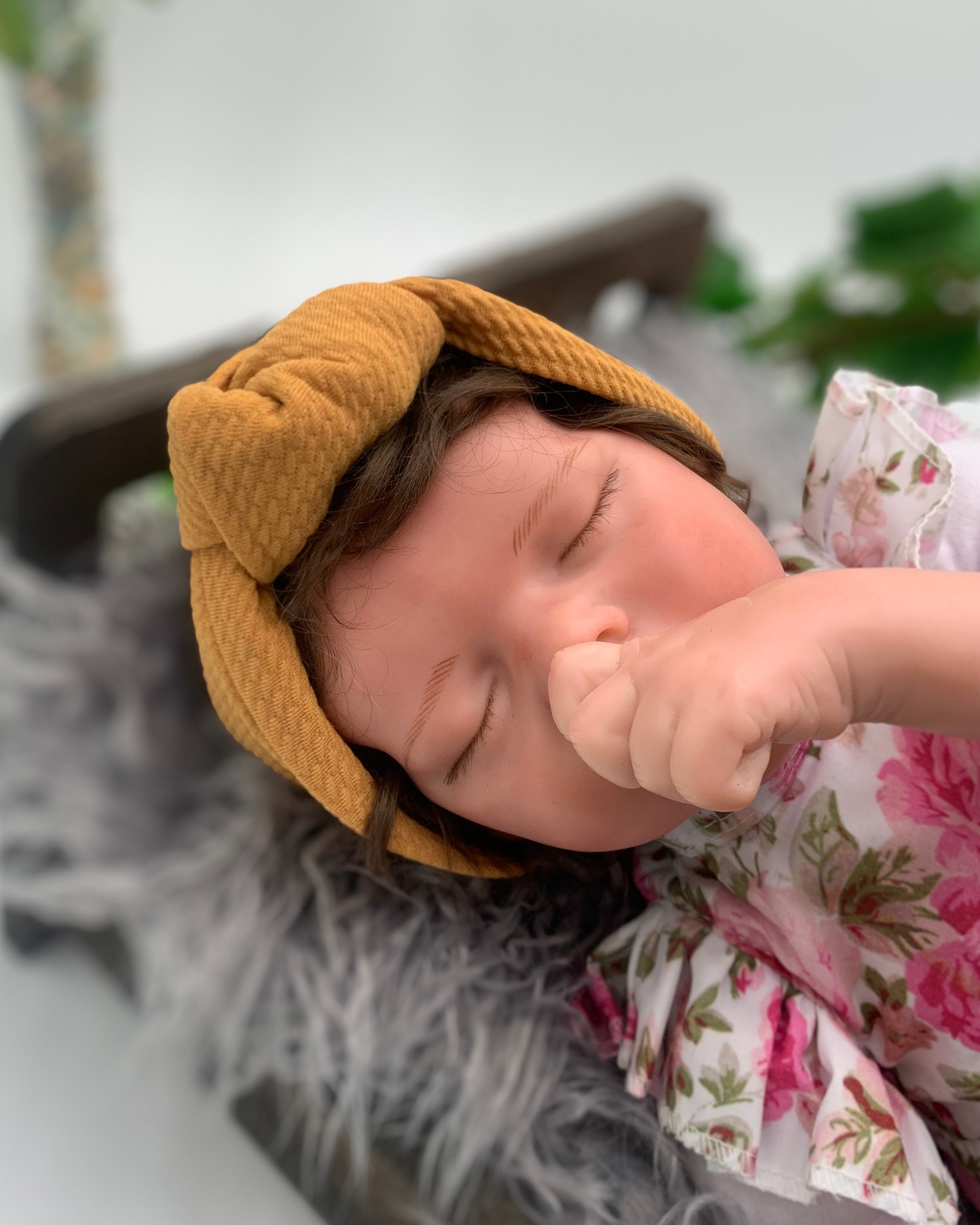 newborn baby doll wearing a Mustard colored knot headband on a white background with green leaves