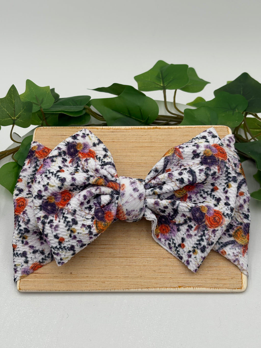 Autumn colored floral themed big bow headband on a wooden board with a white background and greenery 