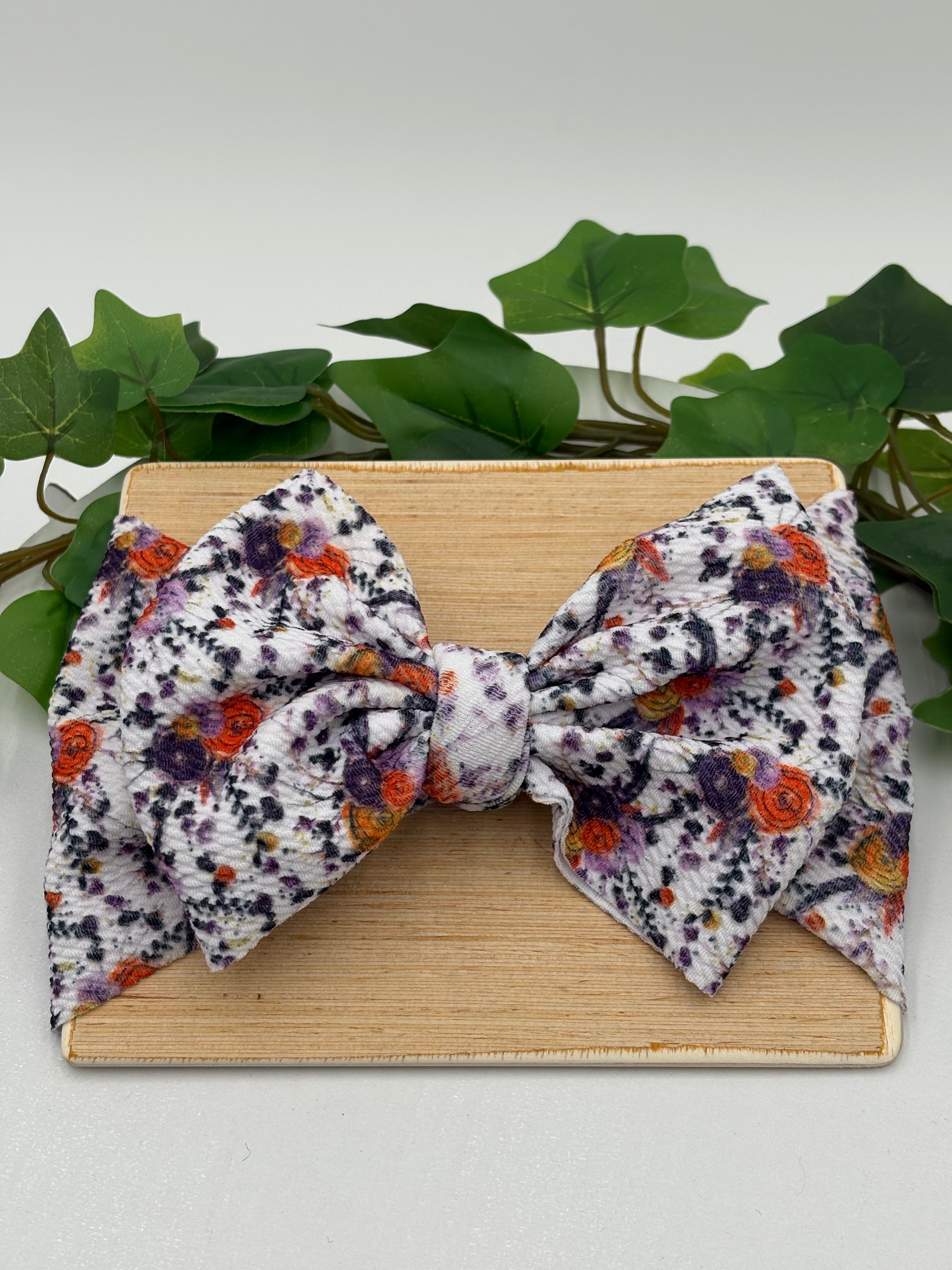 Autumn colored floral themed big bow headband on a wooden board with a white background and greenery 