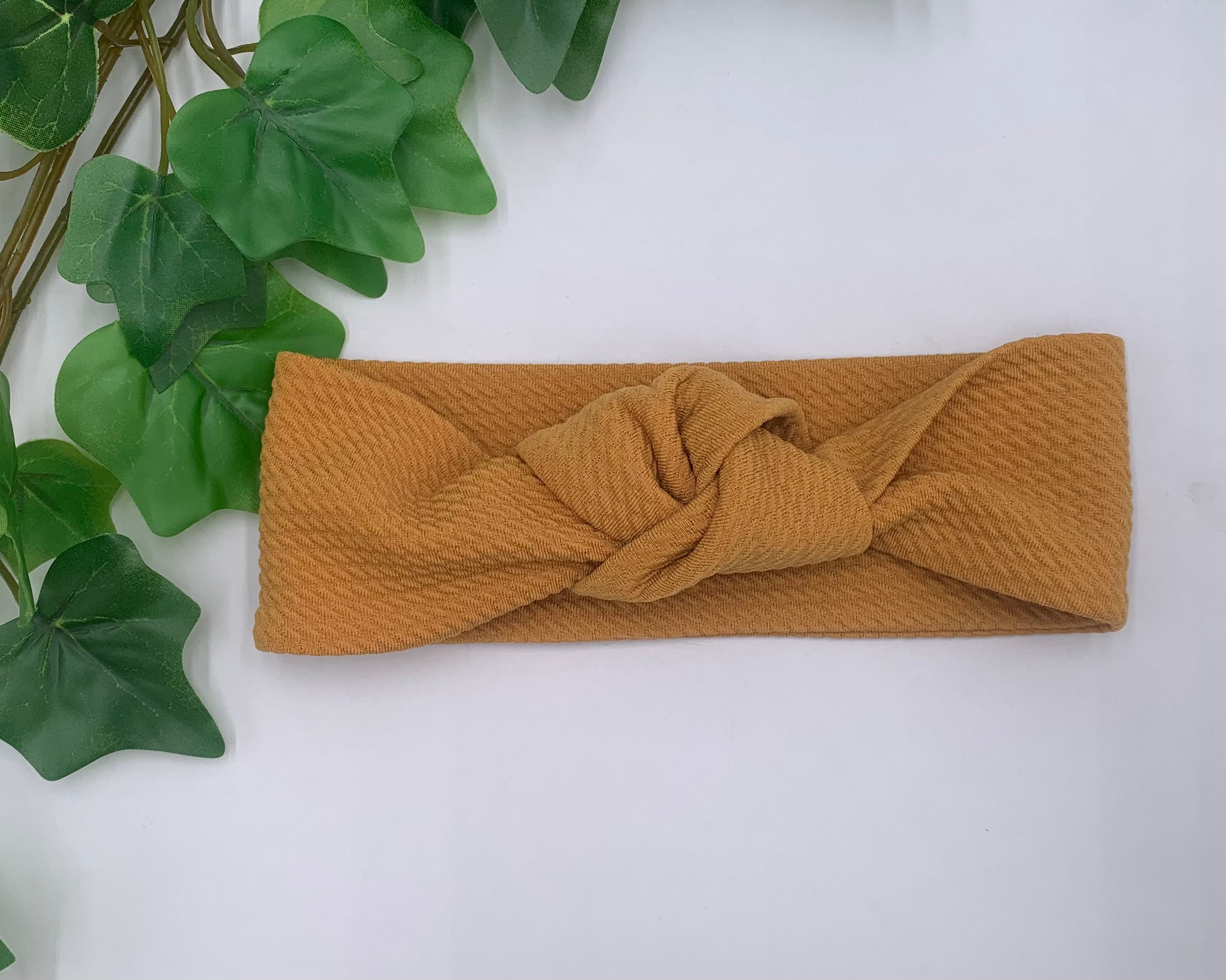 Mustard yellow knotted headband on a white background with green leaves.