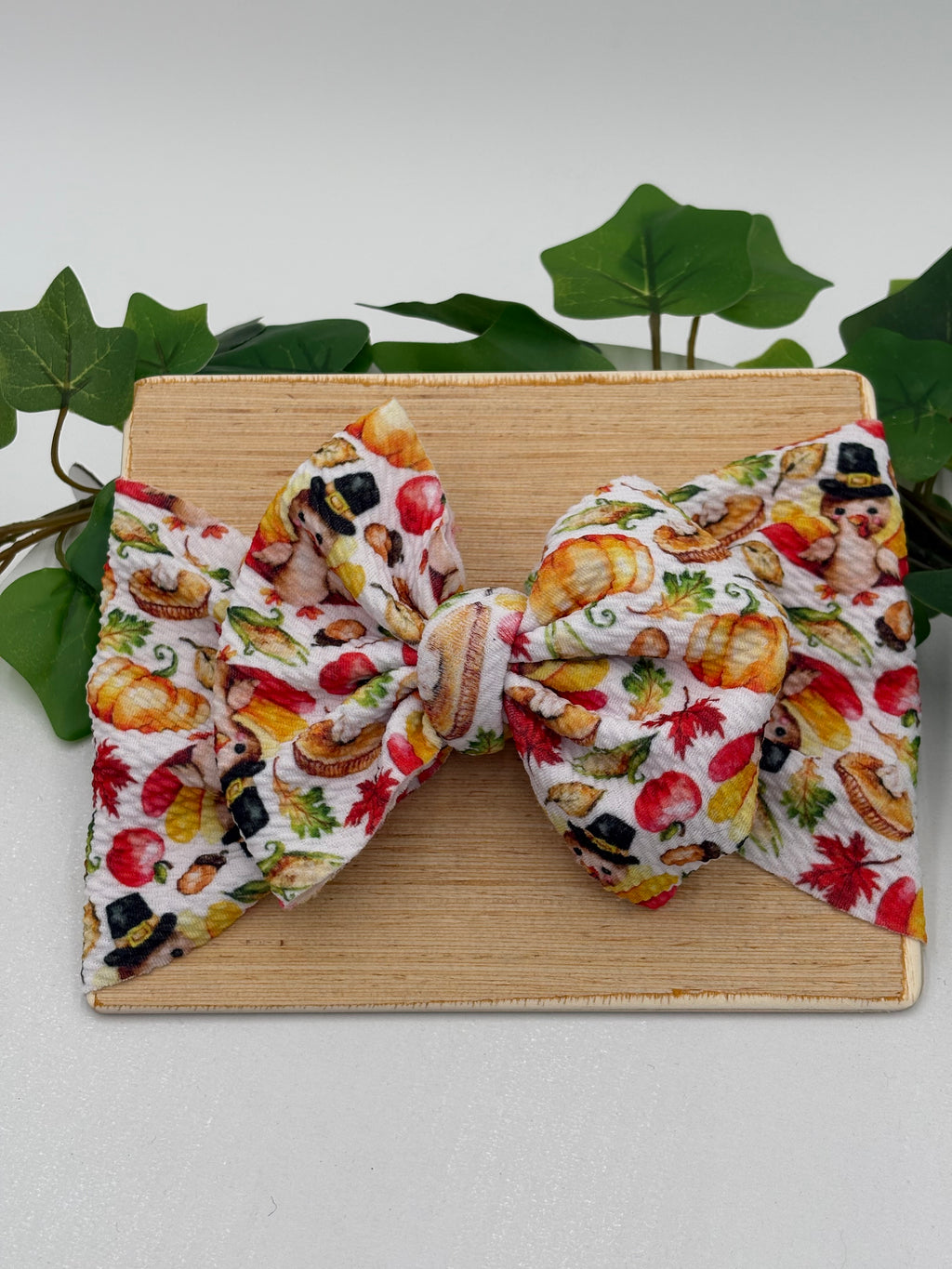 Colorful thanksgiving themed big bow headband on a wooden board with a white background and greenery 
