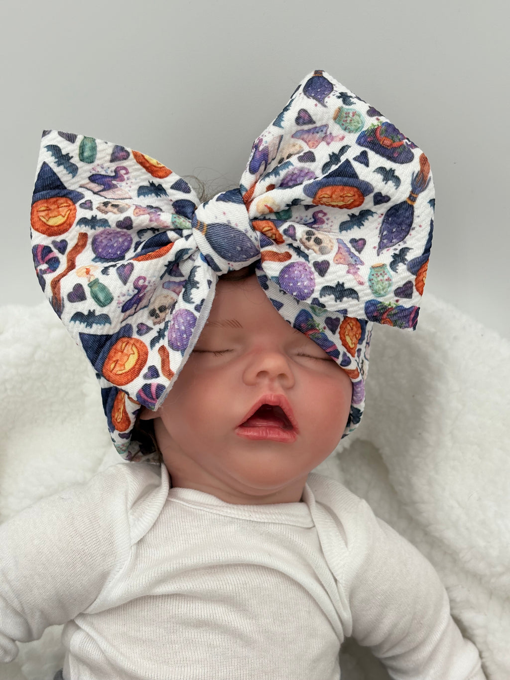 Newborn baby doll wearing a colorful big bow headband with Halloween-themed designs on a white background.