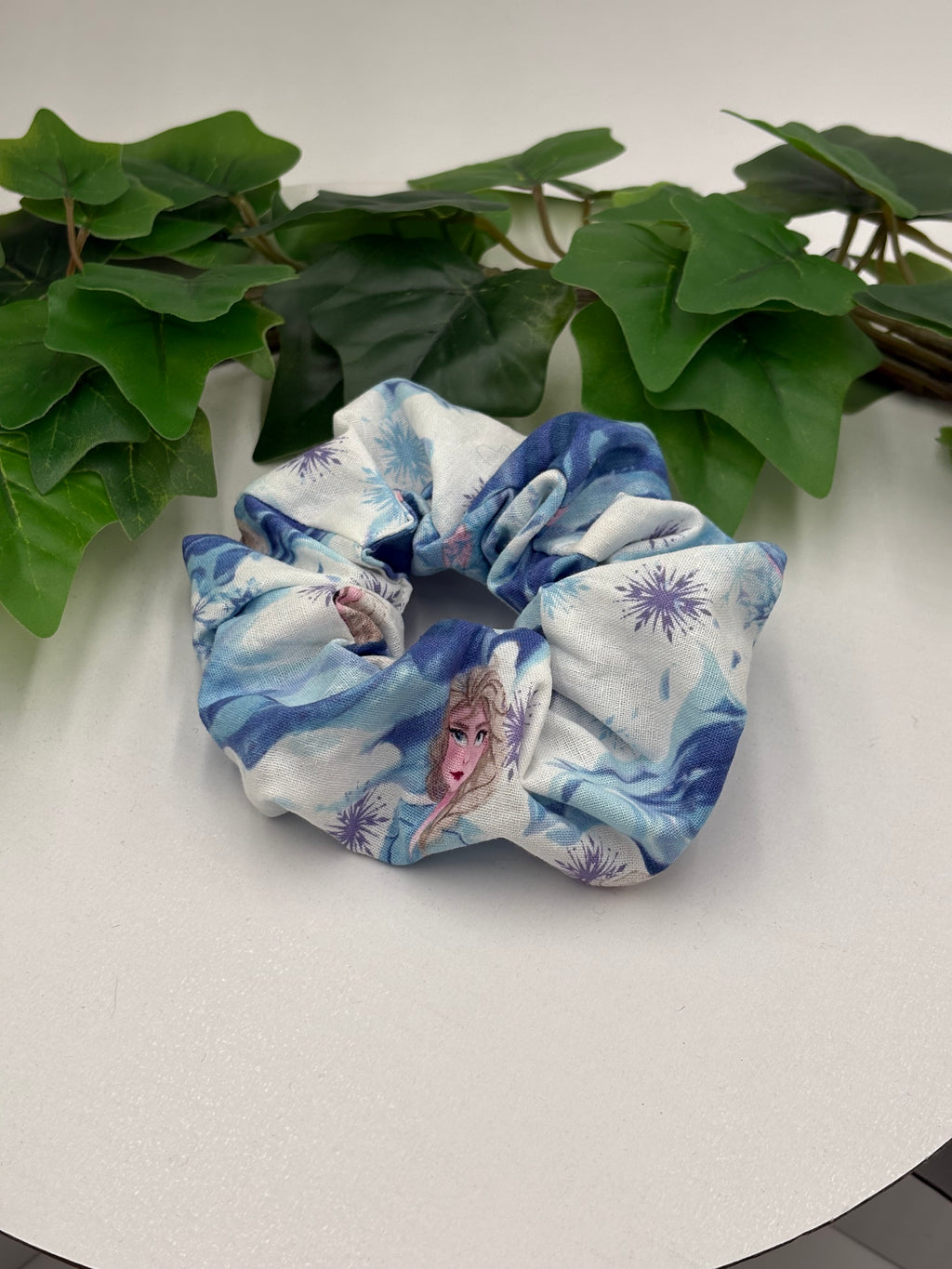 Blue and white scrunchie with character design on a light surface with green leaves.