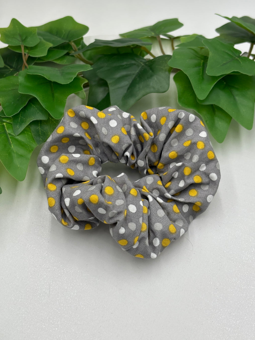 Gray scrunchie with yellow and white polka dots on a light gray background with green leaves.